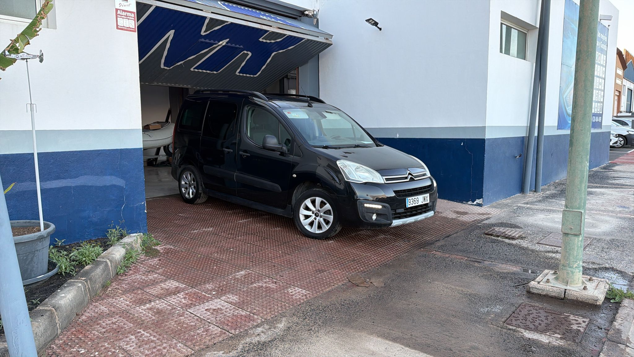 Vehículo Citroën negro estacionado frente a un taller, con un fondo de pared blanca y azul. Se observan detalles del entorno, como una maceta y una señal de alarma.