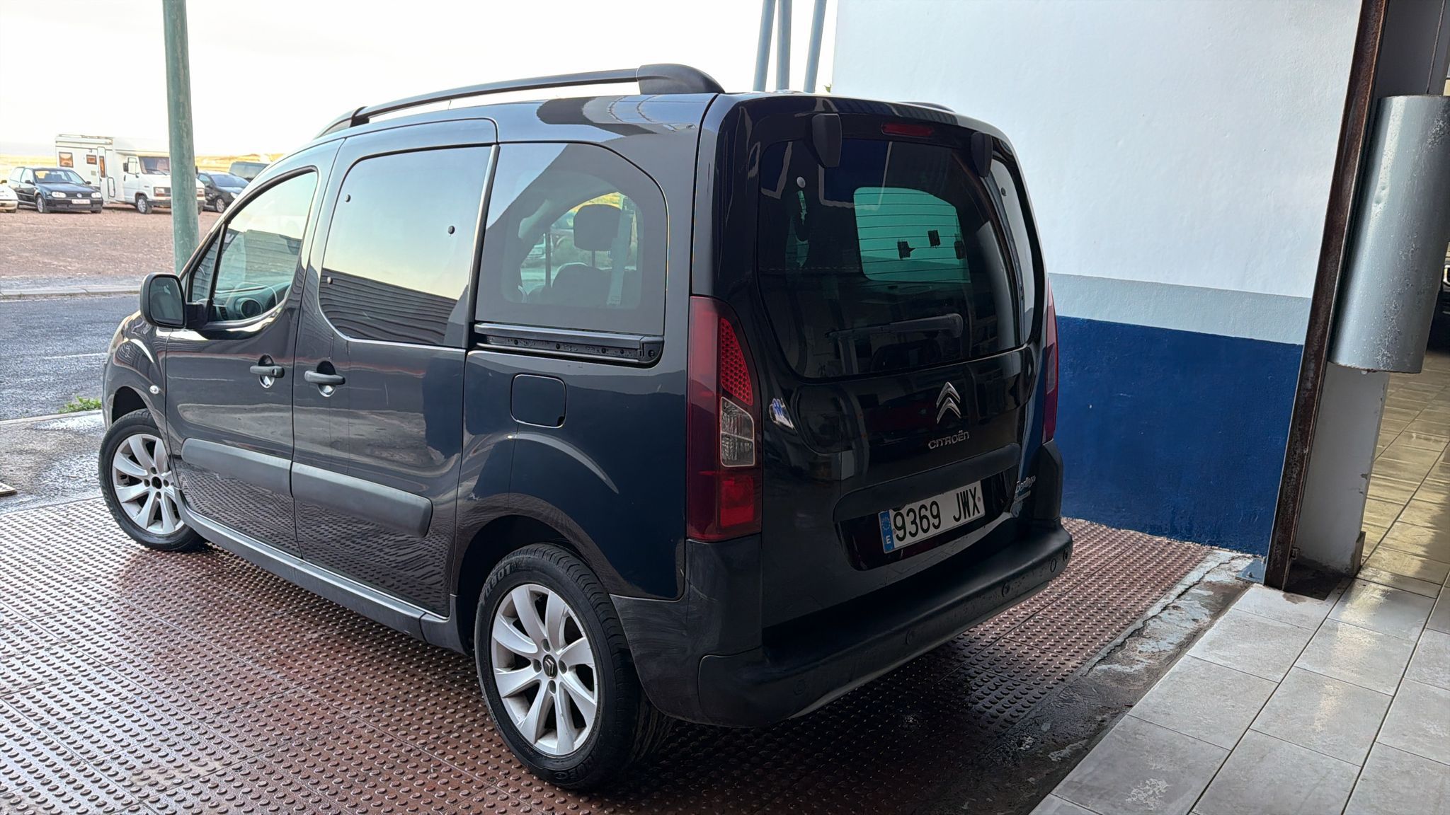 Furgoneta Citroën gris estacionada en un área de carga, mostrando su parte trasera y lateral. El vehículo cuenta con llantas de aleación y un diseño práctico para transporte.