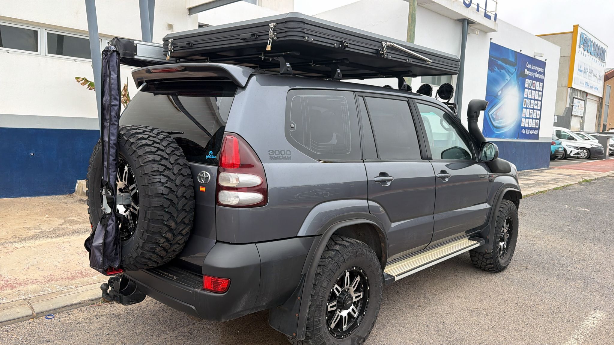SUV Toyota con accesorios para off-road, incluyendo llantas grandes y un toldo en el techo, estacionada frente a un taller mecánico.