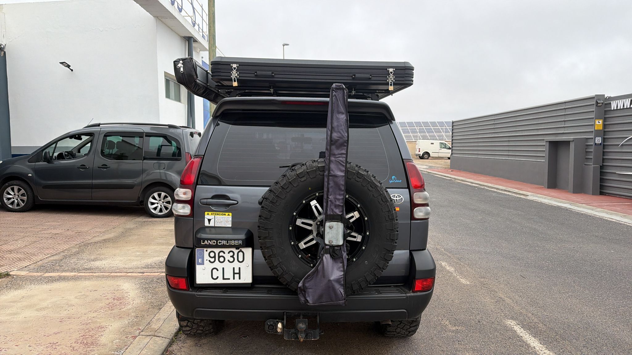 SUV Toyota Land Cruiser estacionada con un portón trasero visible, equipado con una llanta de repuesto y un toldo en el techo. Fondo urbano con otros vehículos y un edificio moderno.