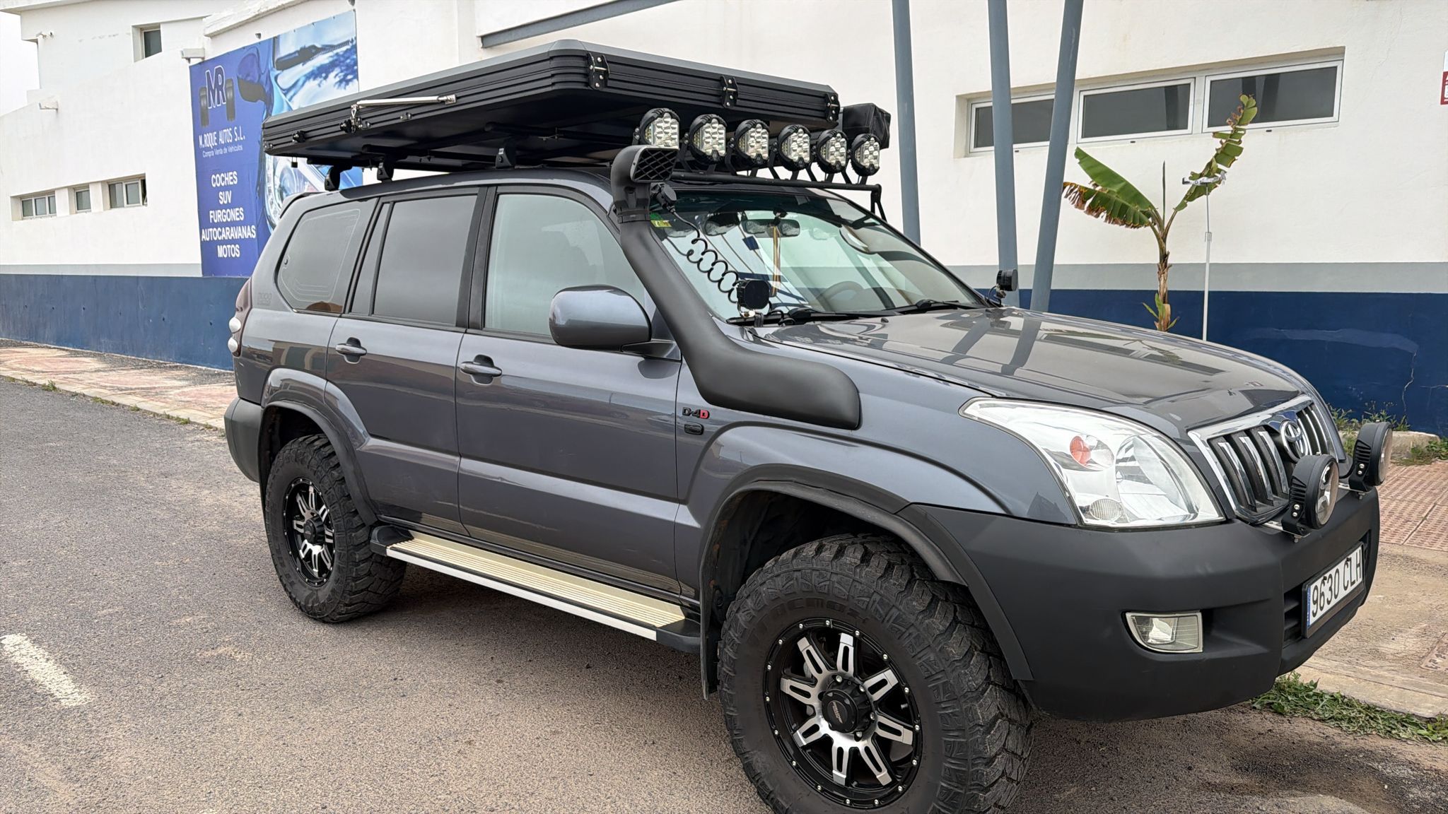 SUV todoterreno gris con accesorios de aventura, como luces LED en el techo y un snorkel, estacionada en una calle. Detrás, se observa un cartel de una empresa de coches y motos.