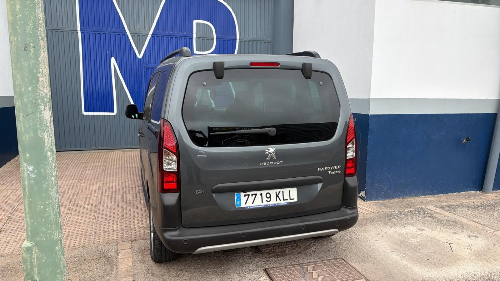 Parte trasera de un Peugeot Partner Tepee gris estacionado frente a una pared con el logo 'M' en color azul. La imagen muestra detalles del vehículo y su matrícula.