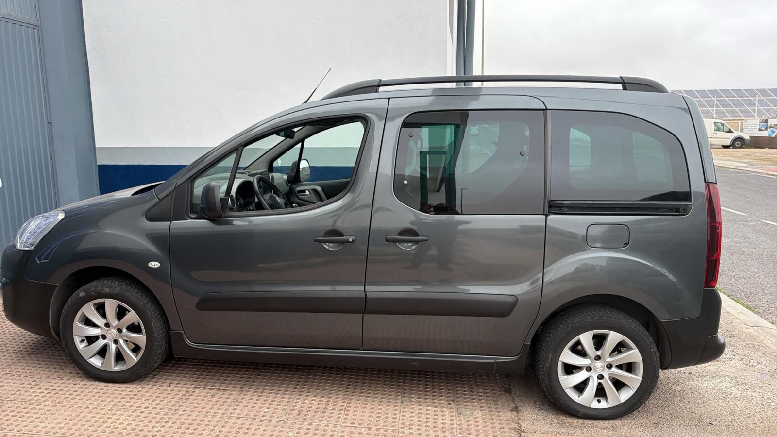 Vehículo familiar gris estacionado, con diseño compacto y amplio interior, ideal para transporte de pasajeros y carga. Se observa en un entorno urbano con un fondo de edificio y espacio abierto.