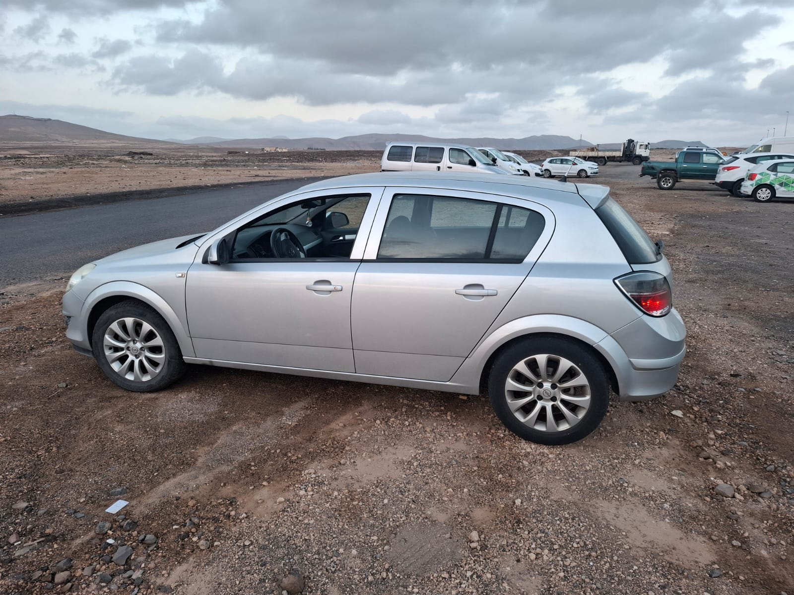Coche compacto plateado estacionado en un terreno de grava, con un paisaje montañoso al fondo y otros vehículos visibles en la distancia.