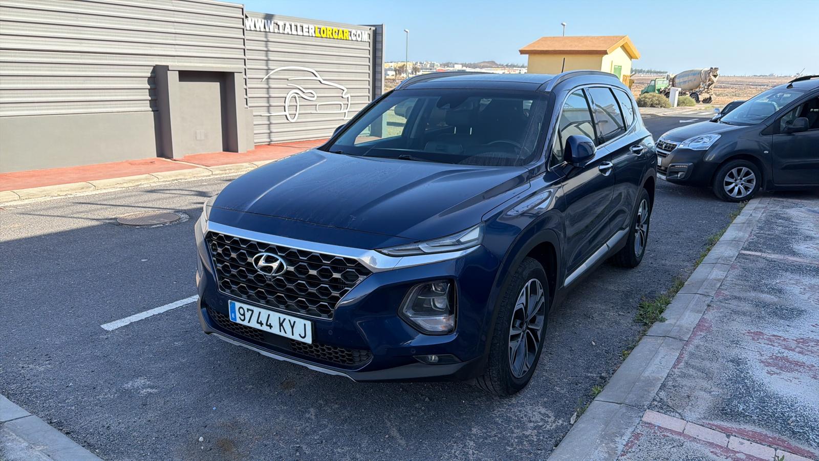 SUV Hyundai de color azul estacionado en la calle, con un diseño moderno y elegante. Se observa el logo de Hyundai en la parrilla frontal y detalles en el acabado. Al fondo, se aprecian otros vehículos y un taller de automóviles.