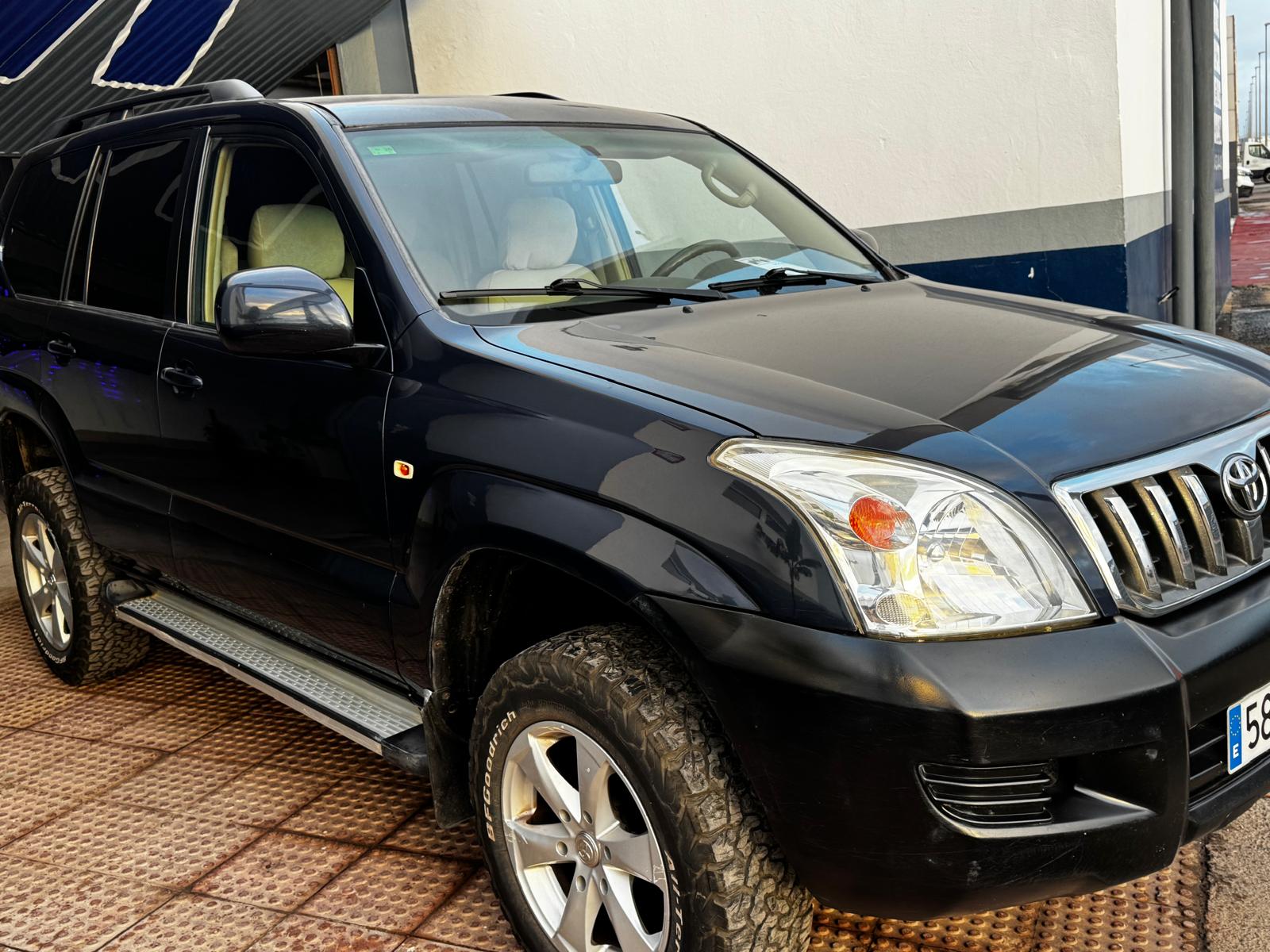 SUV Toyota negra estacionada, con llantas robustas y detalles cromados, frente a un fondo de pared blanca y un área de estacionamiento. Ideal para aventuras off-road y conducción urbana.