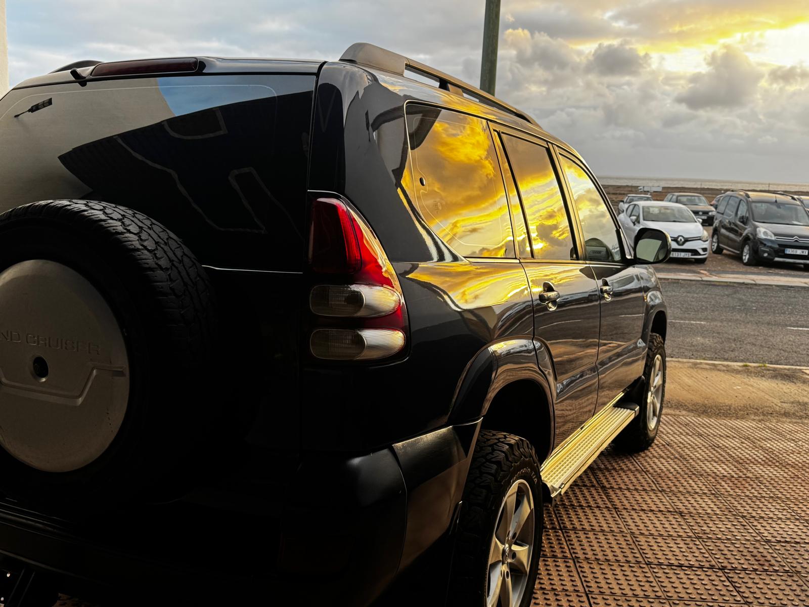 SUV negra estacionada con reflejos del atardecer en las ventanas, mostrando el paisaje costero y otros vehículos de fondo. Ideal para resaltar características de diseño y versatilidad.