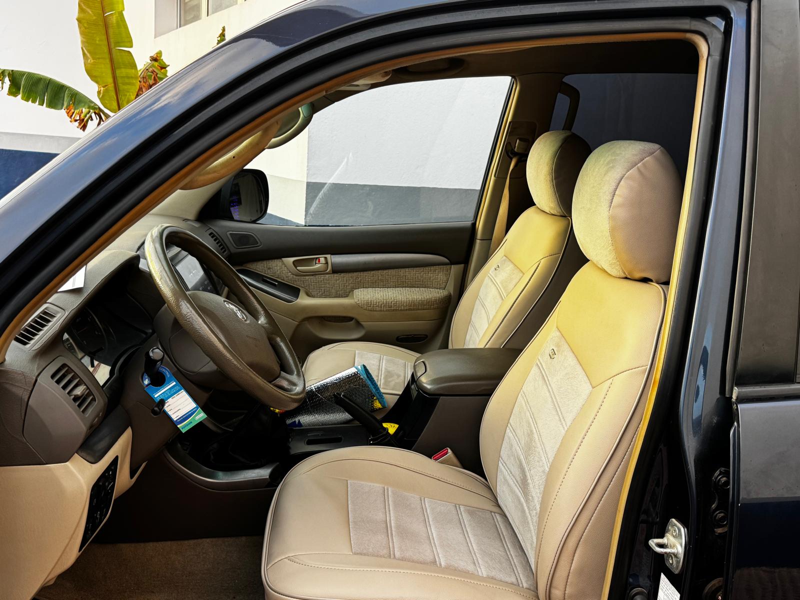 Interior de un automóvil con asientos de cuero beige y un volante de cuero. Se observa un espacio organizado con documentos y un espejo retrovisor. Fondo con una pared blanca y una planta visible.