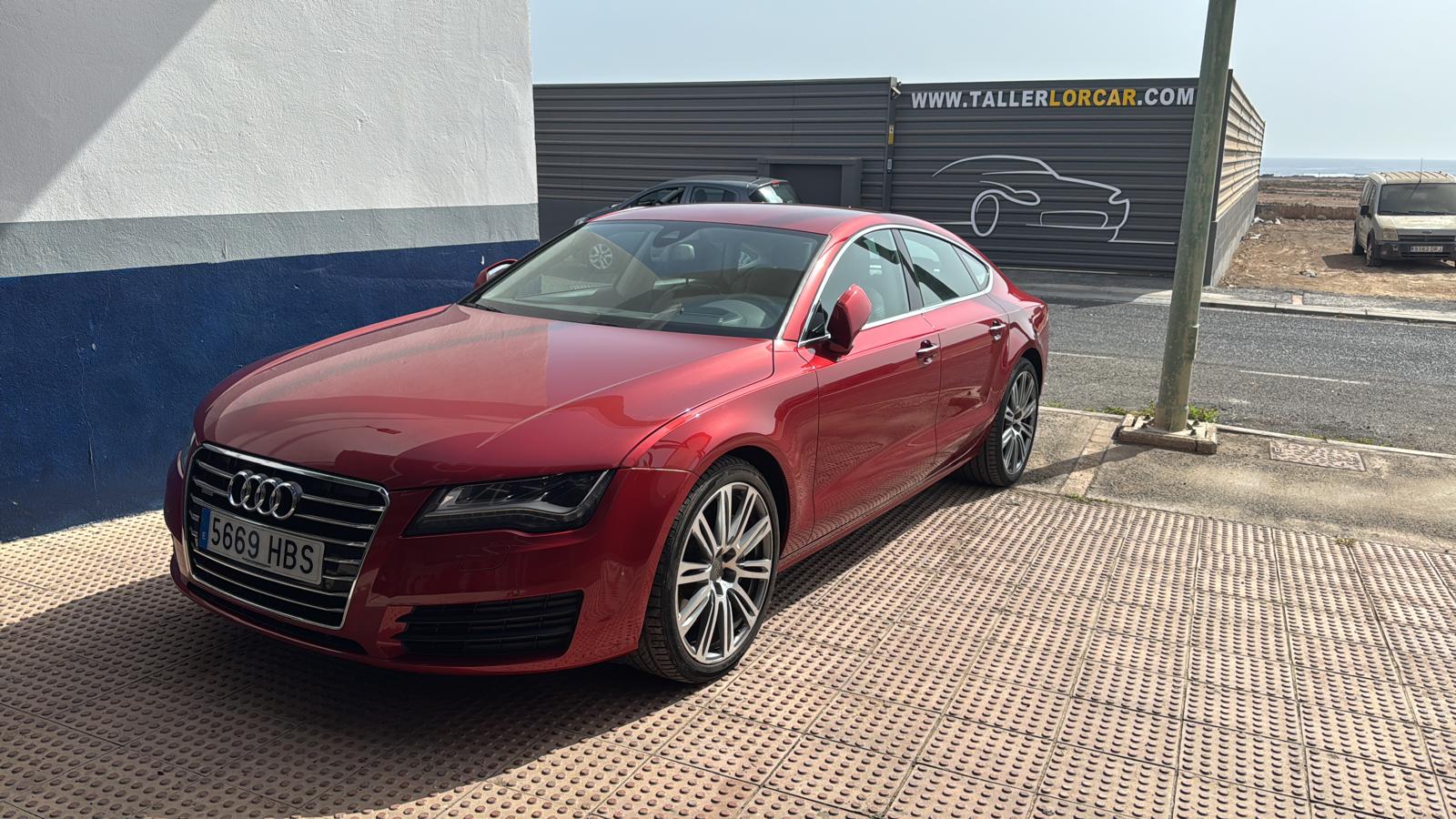 Audi A7 de color rojo estacionado frente a un taller mecánico, con un diseño elegante y moderno, destacando sus llantas de aleación y detalles distintivos de la marca.
