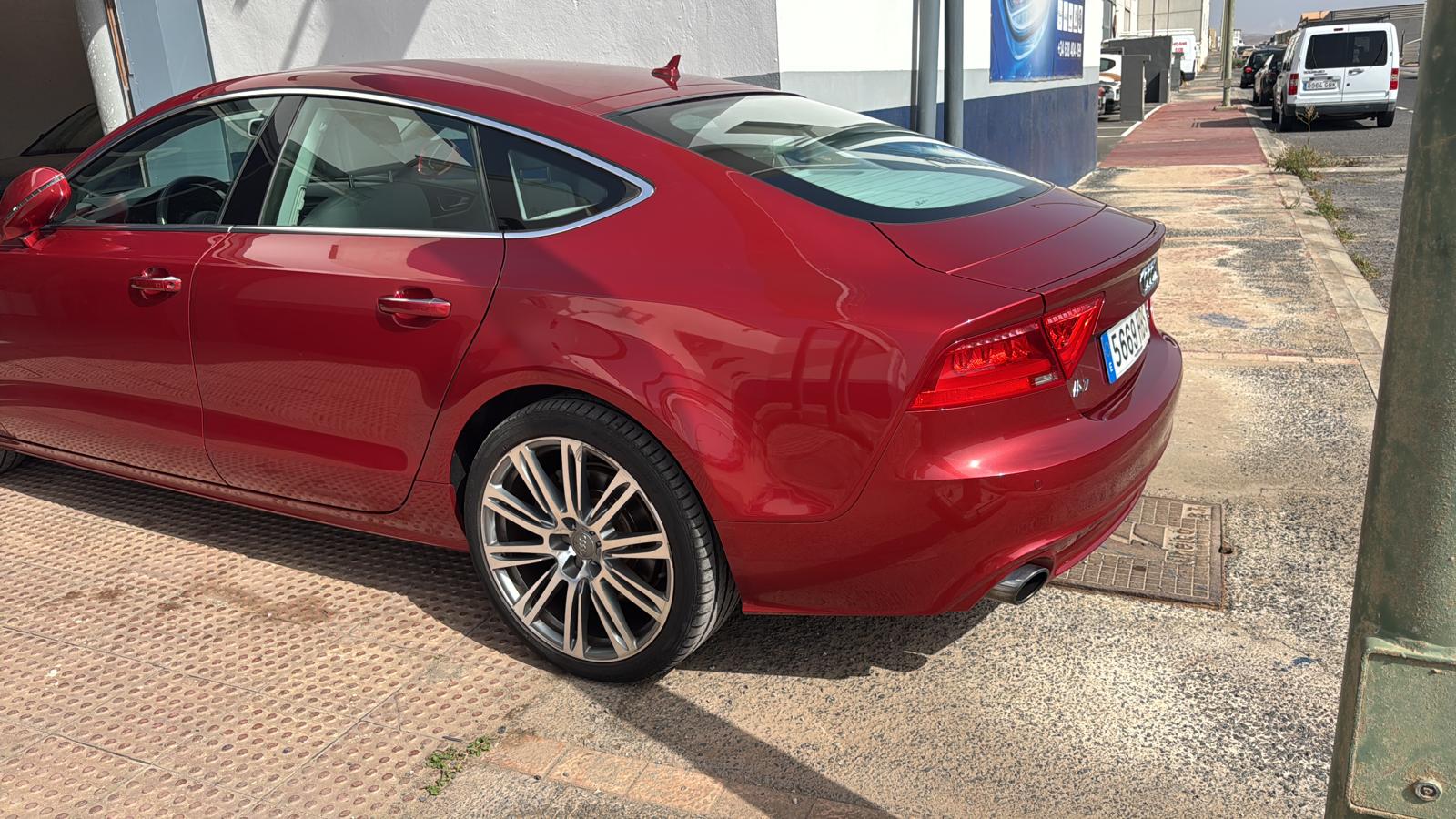 Audi rojo estacionado en la calle, mostrando su diseño elegante y llantas de aleación. El vehículo está parcialmente cubierto por sombra, destacando su acabado brillante y detalles de estilo.