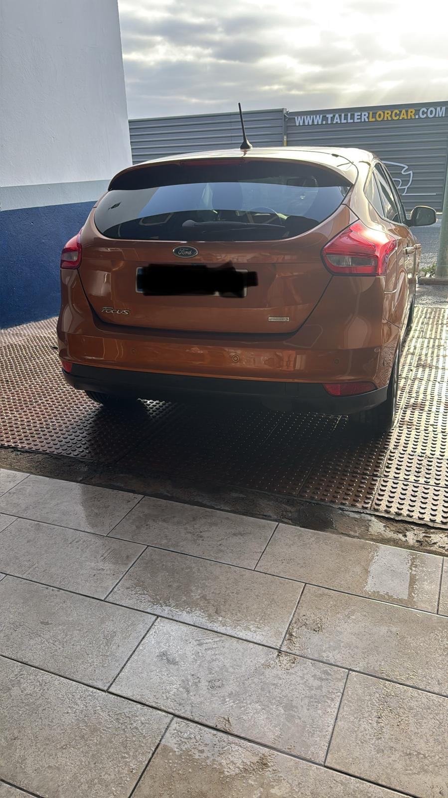 Ford Focus de color naranja estacionado en un taller, con un fondo de cielo nublado y piso de baldosas.