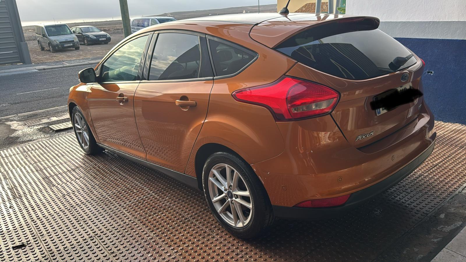 Ford Focus naranja estacionado en una superficie metálica, con vista lateral y trasera, en un entorno urbano con otros vehículos al fondo.