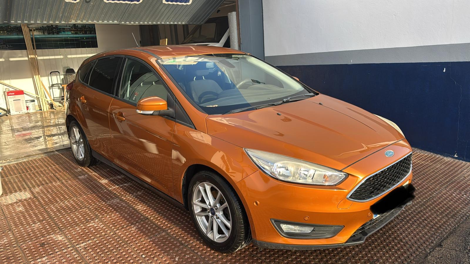 Ford Focus de color naranja brillante, estacionado en un lavado de autos, con llantas de aleación y un diseño moderno. El vehículo está en buen estado y presenta un acabado limpio.