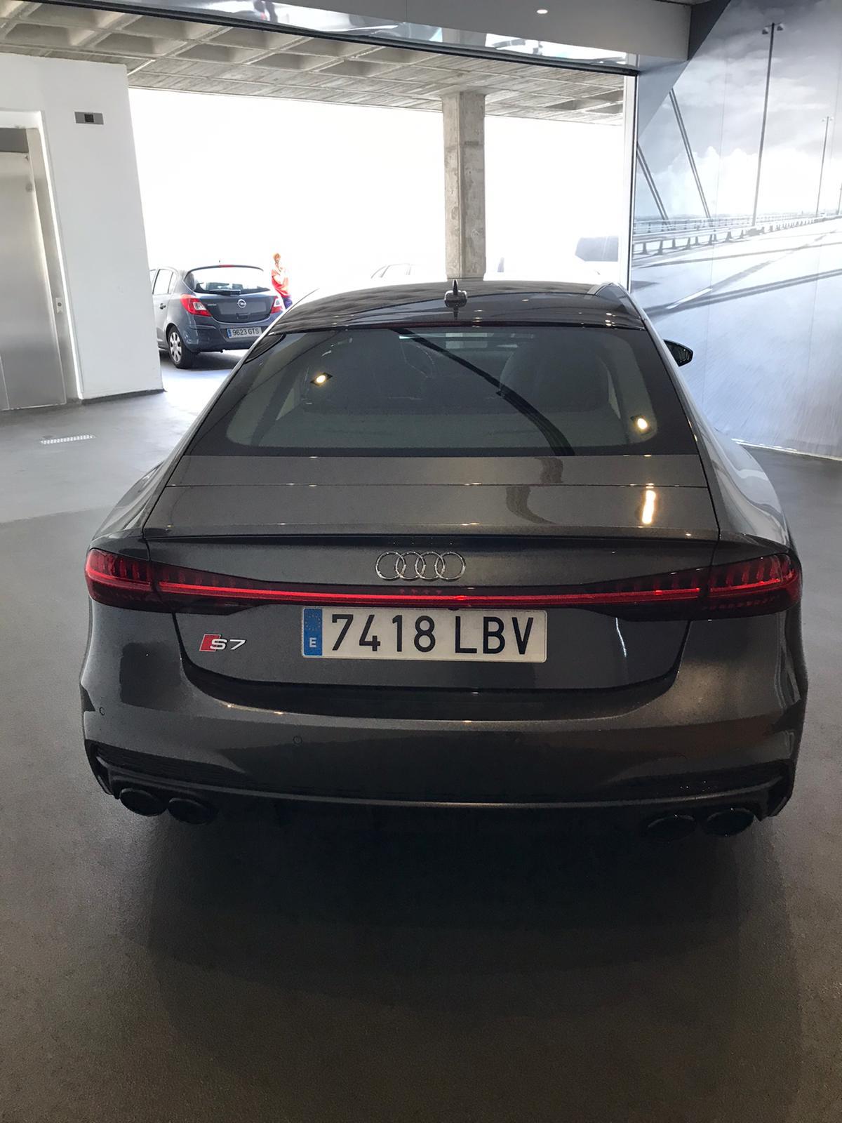 Audi S7 gris aparcado en un garaje, mostrando su parte trasera y placa de matrícula 7418 LBV. El ambiente es moderno y luminoso, con un fondo de diseño arquitectónico.