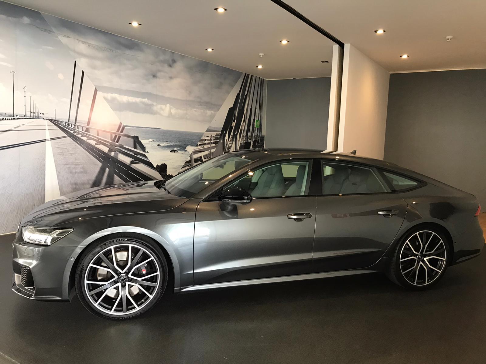 Automóvil Audi A7 Sportback de color gris oscuro, exhibido en un showroom moderno con fondo de paisaje costero y carretera. El vehículo destaca por sus llantas de diseño deportivo y su elegante línea.