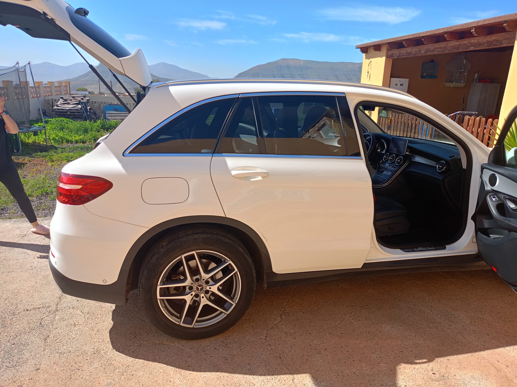 SUV blanca Mercedes-Benz GLC estacionada al aire libre, con el maletero abierto y la puerta del conductor abierta, mostrando el interior moderno y espacioso. Fondo con montañas y un paisaje verde.
