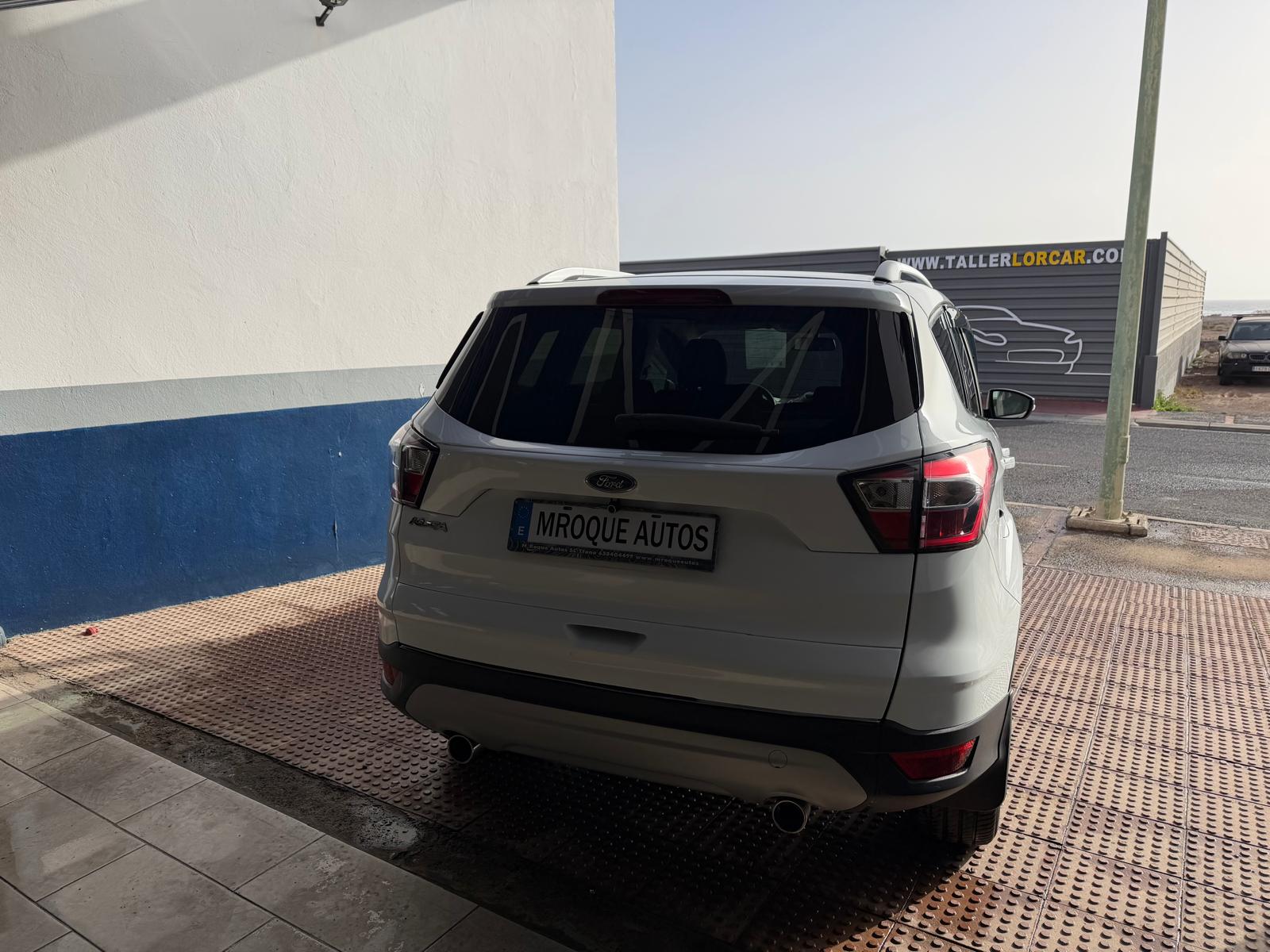 SUV Ford blanca estacionada en un taller de autos, con un fondo que muestra una pared blanca y azul. El letrero del taller es visible en el fondo, indicando servicios de reparación de vehículos.