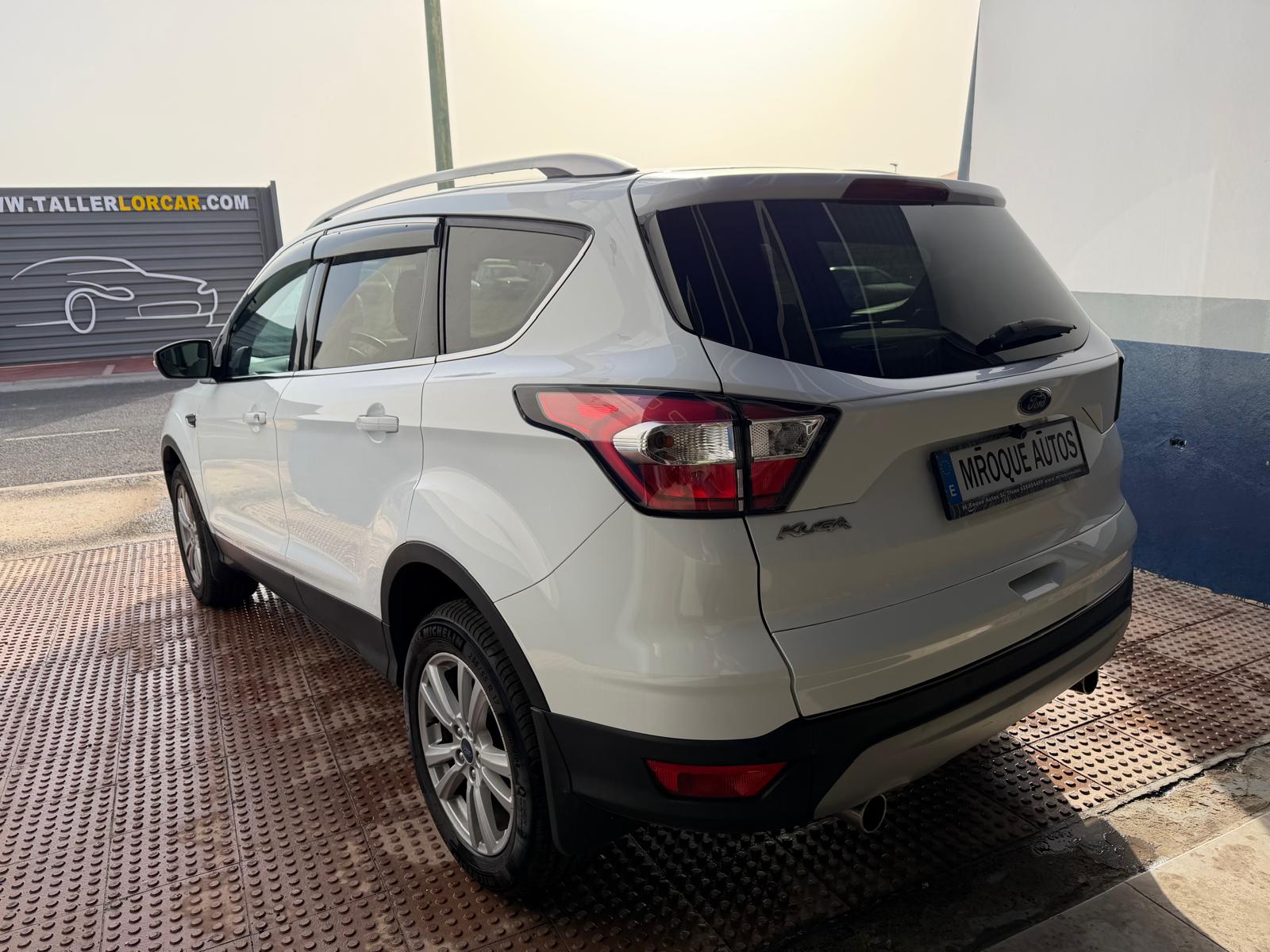 SUV Ford Kuga blanca estacionada en un taller, mostrando detalles del diseño trasero y lateral, con un fondo de un letrero de taller automotriz.