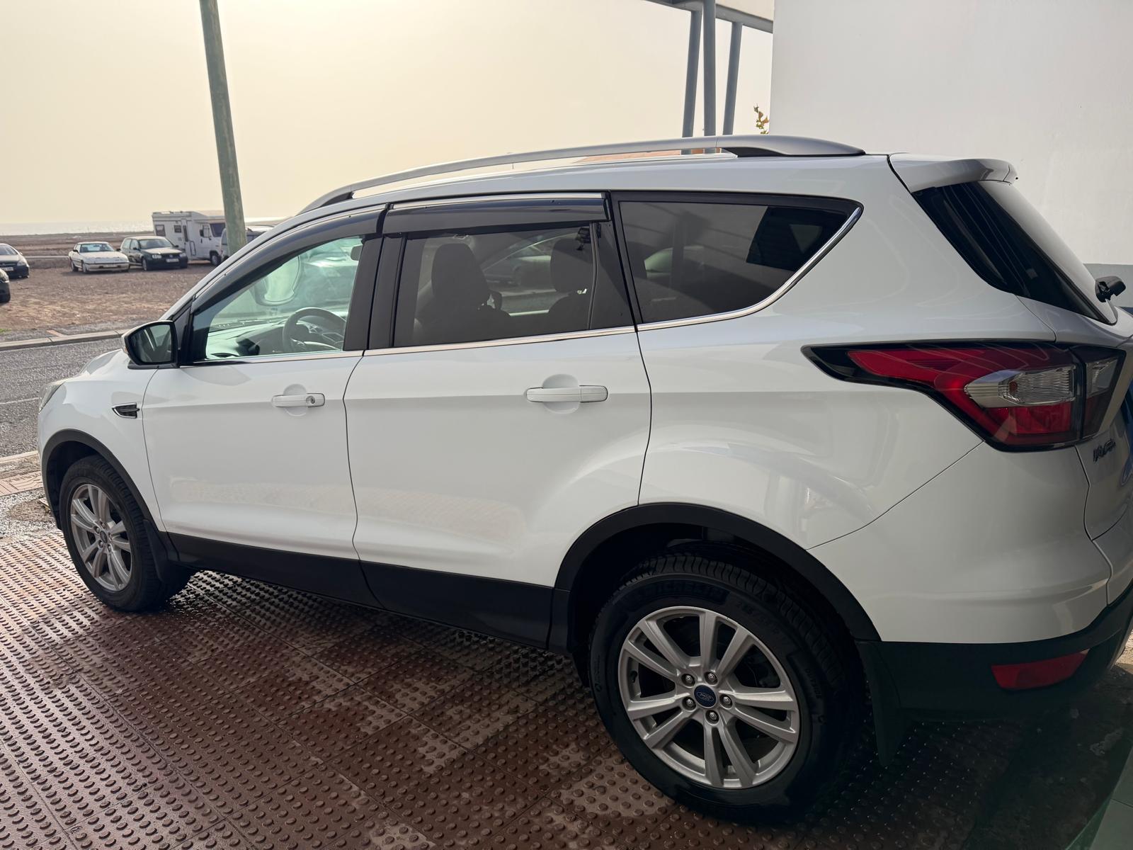 SUV blanca estacionada en un área con pavimento de goma, mostrando su diseño moderno y llantas de aleación. Al fondo, se pueden ver otros vehículos y un paisaje costero difuso.