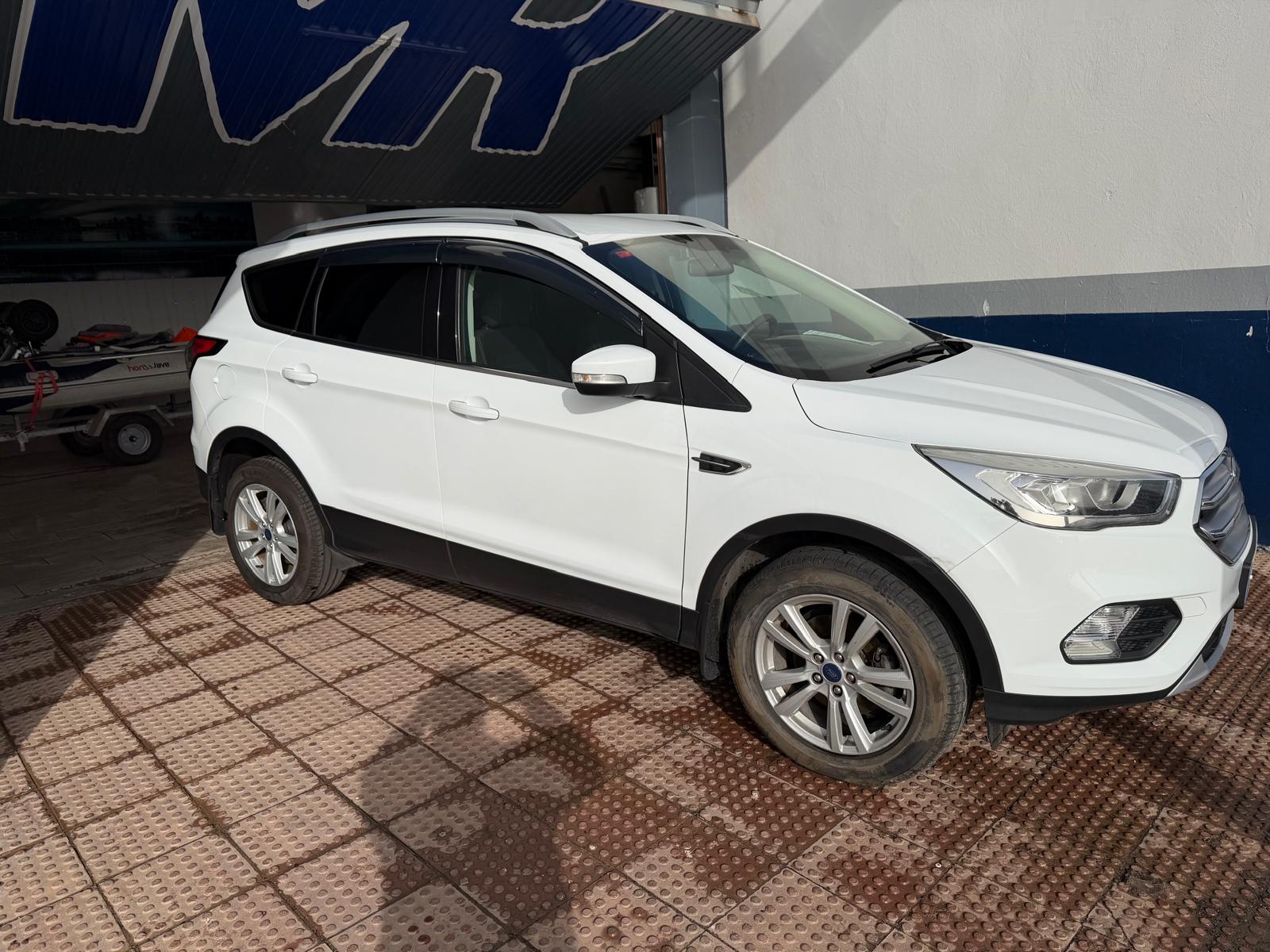 SUV blanca Ford Edge estacionada sobre un pavimento de loseta, con un fondo que muestra una estructura de taller. Ideal para destacar características de vehículos y su diseño moderno.