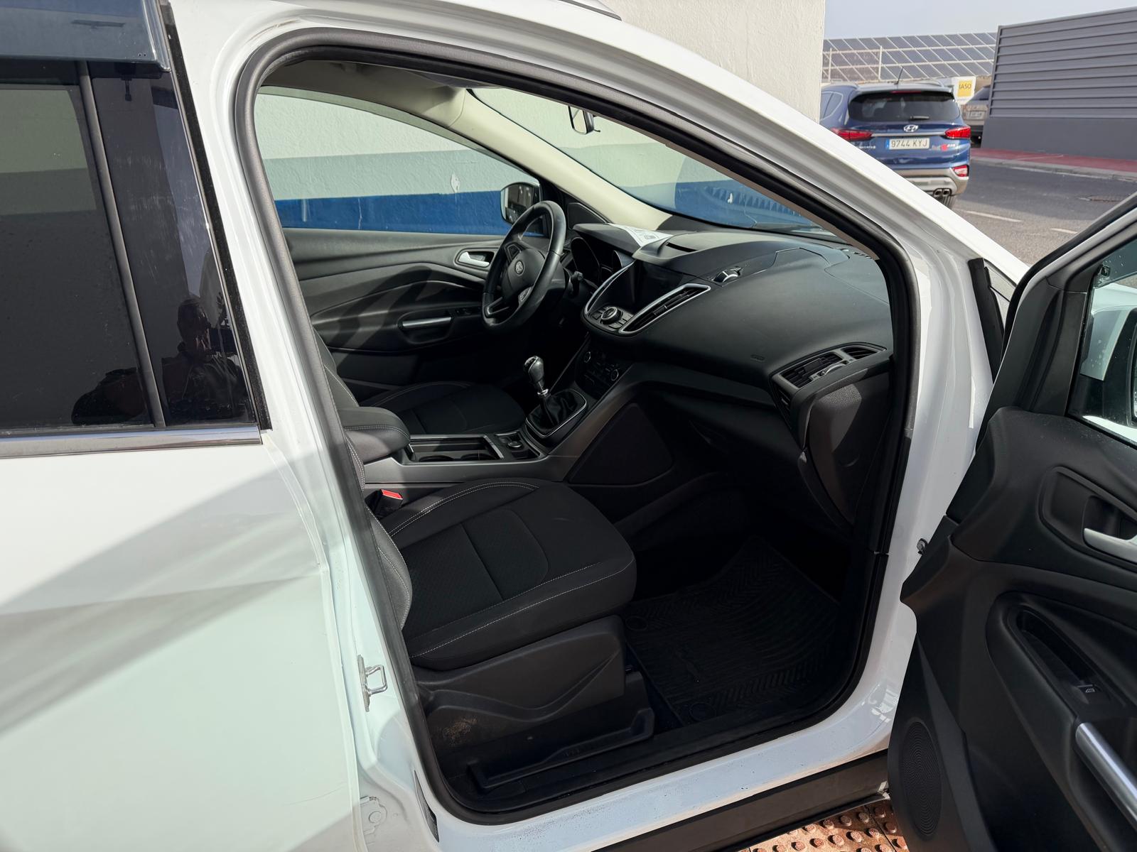 Interior de un automóvil blanco, mostrando el asiento del conductor y el panel de control. Se observa una cabina moderna y espaciosa, con asientos de tela negra y detalles en el tablero. La puerta del lado del conductor está abierta, ofreciendo una vista amplia del interior.