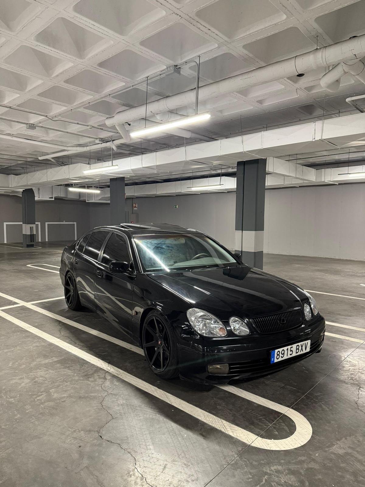 Automóvil negro estacionado en un garaje, con llantas de diseño deportivo y detalles estilizados. Fondo de concreto y techos con iluminación LED.