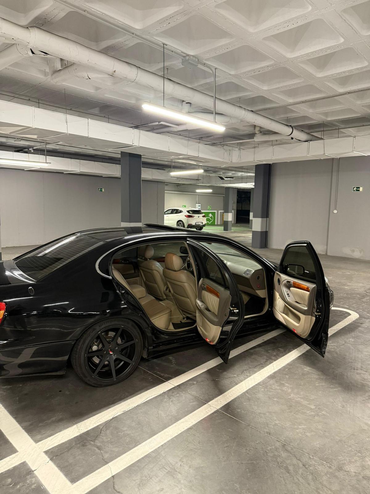 Automóvil negro estacionado en un garaje, con las puertas abiertas y asientos de cuero beige visibles. El entorno muestra un espacio de estacionamiento moderno y bien iluminado.