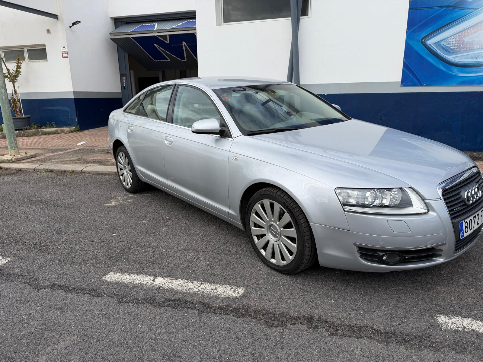 Audi A6 plateado estacionado en la calle, con un edificio de fondo que presenta un diseño moderno. El vehículo muestra un perfil elegante y llantas de aleación.