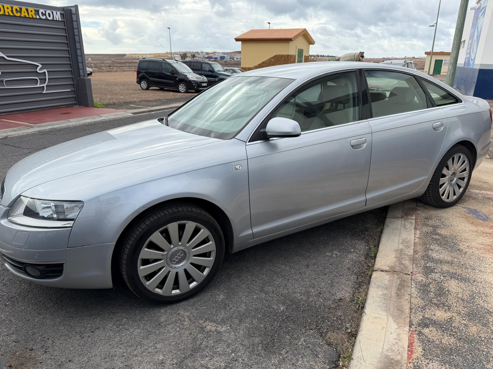 Audi A6 plateado estacionado en una calle, con llantas de diseño elegante y un fondo que muestra un edificio y vehículos. Ideal para promover alquiler de coches o venta de vehículos de lujo.