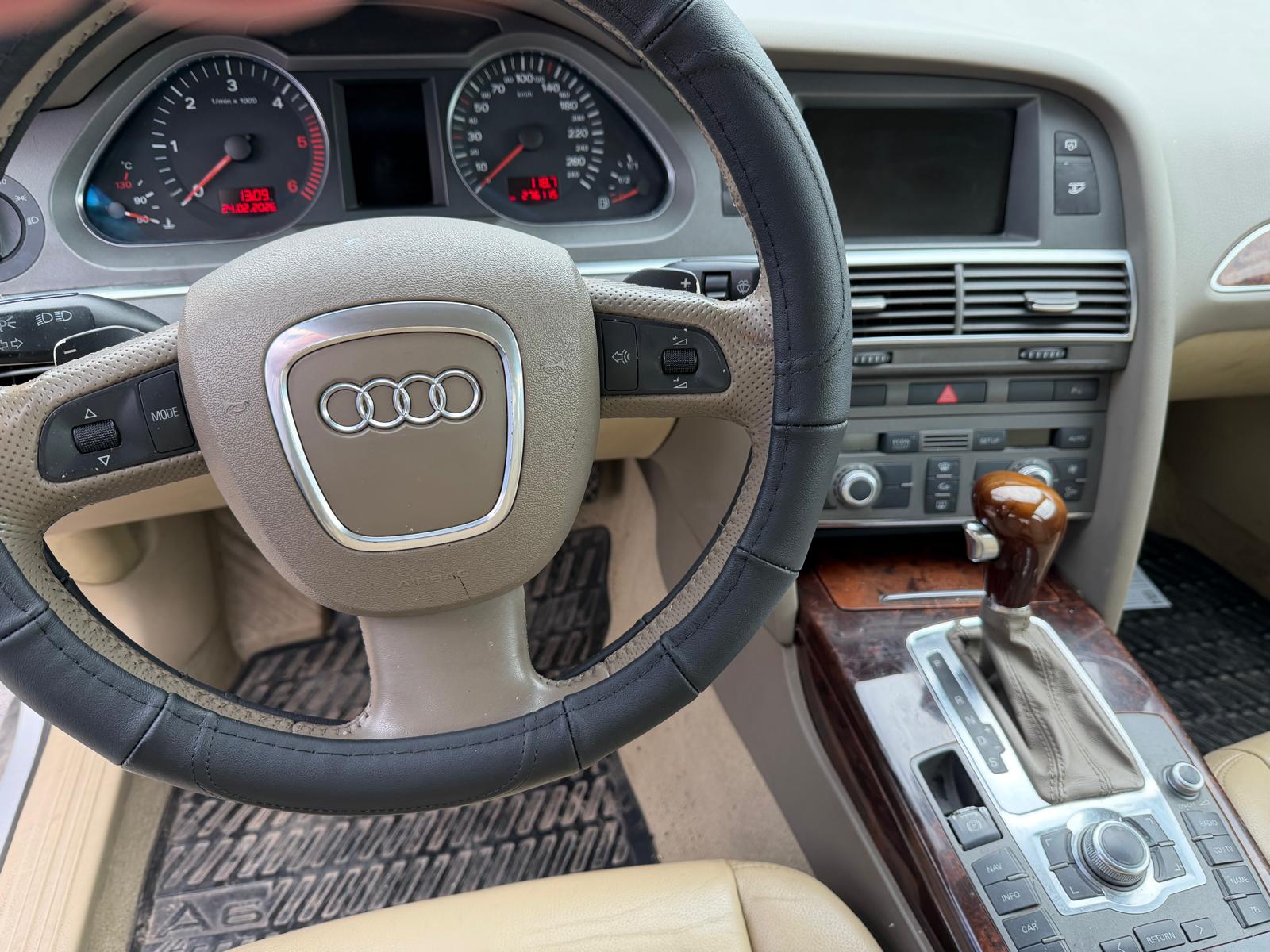 Interior de un Audi A6, mostrando el volante, el tablero de instrumentos con indicadores digitales y una consola central con controles de audio y transmisión automática.