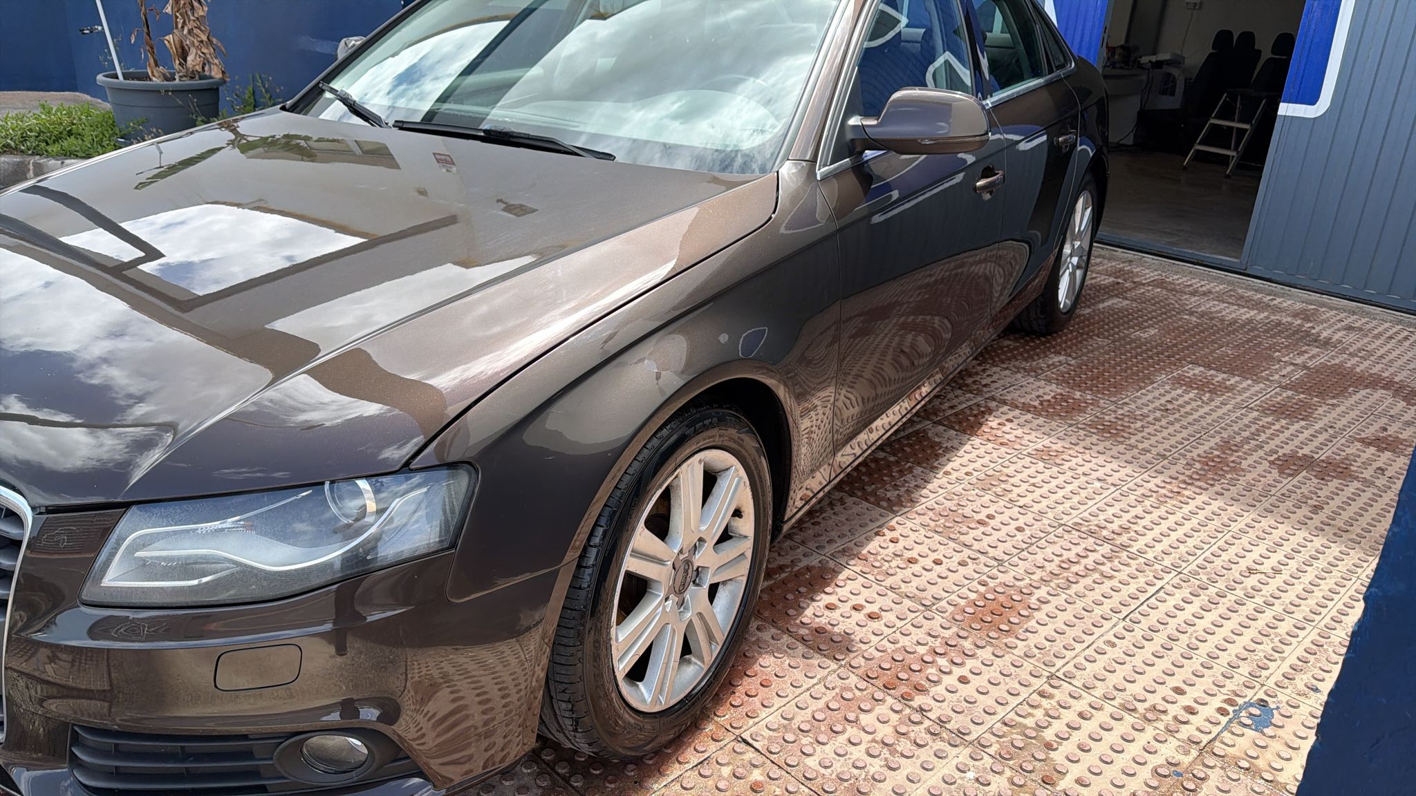 Coche Audi A4 de color marrón estacionado en una superficie de piso metálico, con reflejos del cielo en la carrocería y un fondo de taller visible.