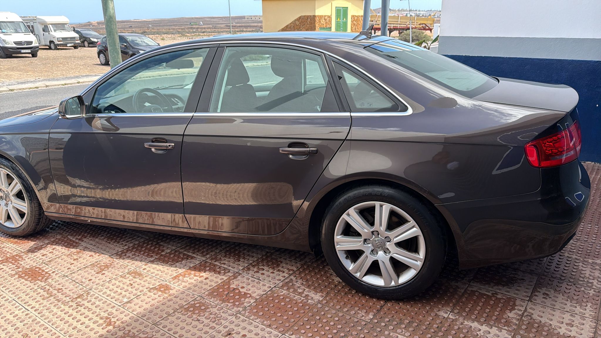 Automóvil de color marrón estacionado en una superficie de baldosas, mostrando su perfil lateral y llantas de aleación. Al fondo, se pueden ver otros vehículos y un paisaje árido.