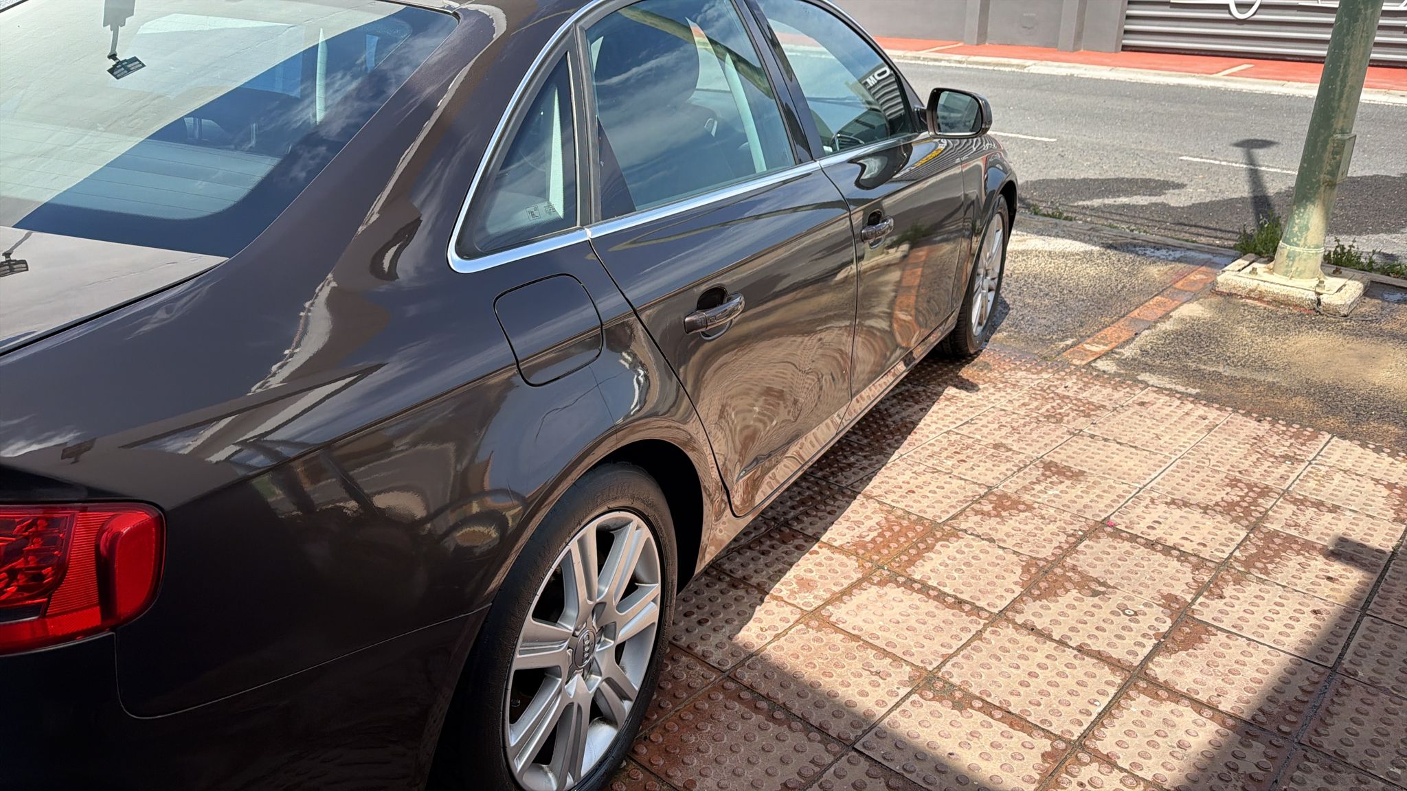 Automóvil de color marrón estacionado sobre un pavimento de ladrillos, mostrando su diseño elegante y llantas de aleación. Reflejos de luz sobre la carrocería y un entorno urbano de fondo.