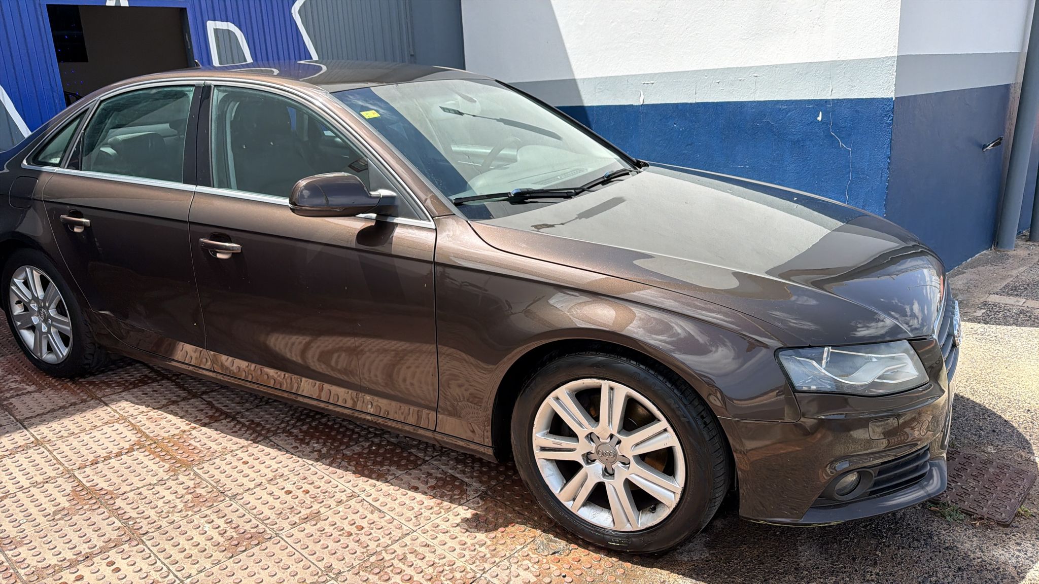 Automóvil Audi A4 color marrón estacionado en un área pavimentada, con llantas de aleación y un fondo de pared azul y blanca. Ideal para promocionar coches de lujo o servicios de venta de vehículos.