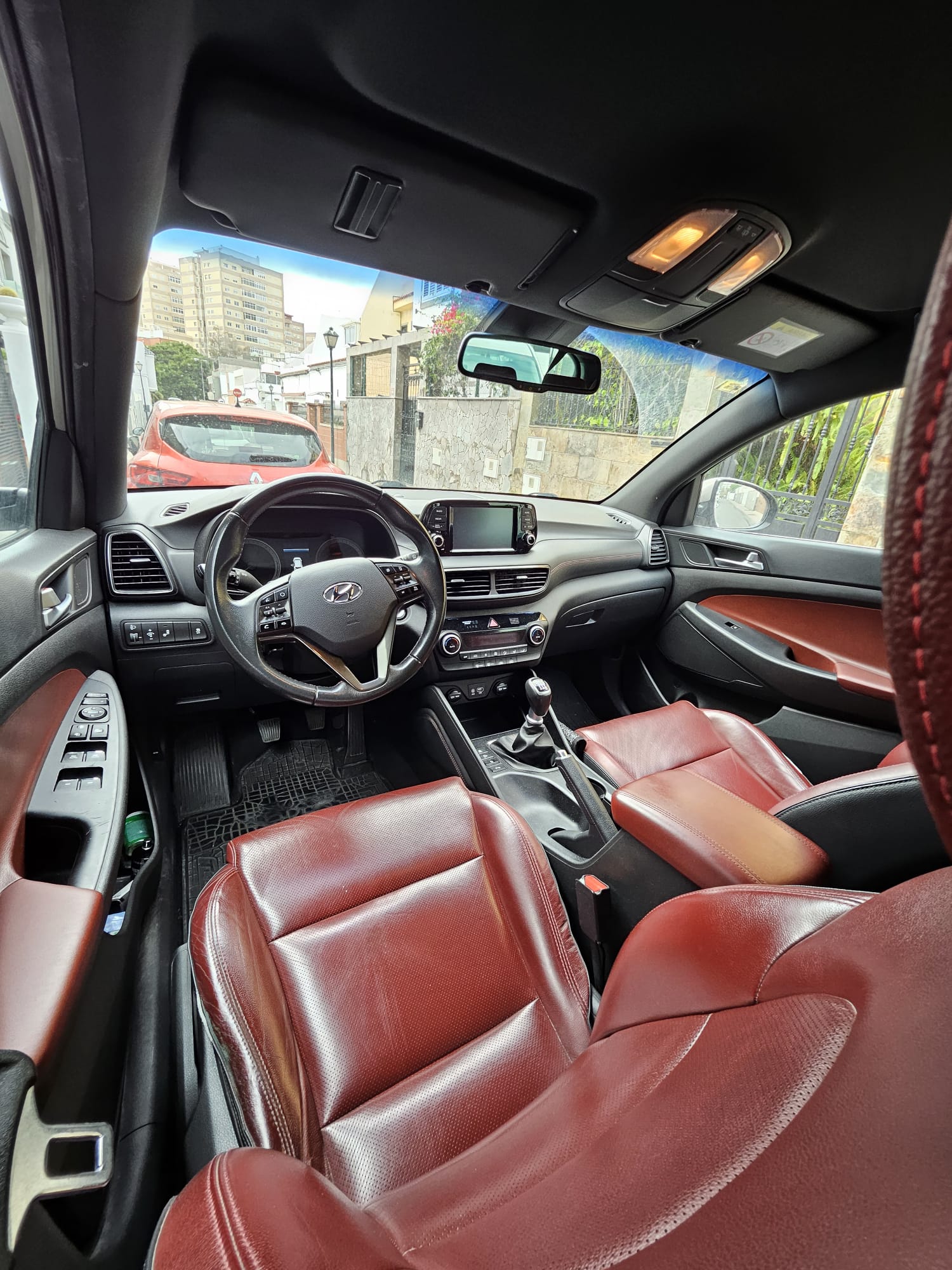 Interior de un automóvil Hyundai con asientos de cuero rojo, vista desde el asiento del conductor, mostrando el volante, la consola central y el tablero, en un entorno urbano.