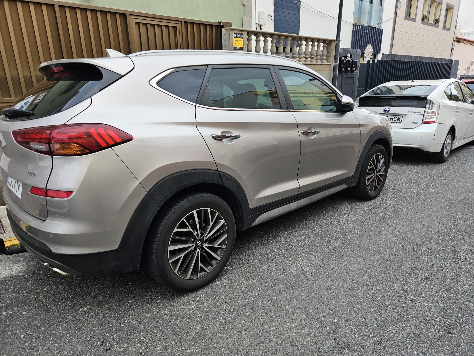 SUV plateado aparcado en la calle, con llantas de diseño moderno y luces traseras distintivas, junto a un vehículo blanco. Fondo urbano con edificios y pavimento.