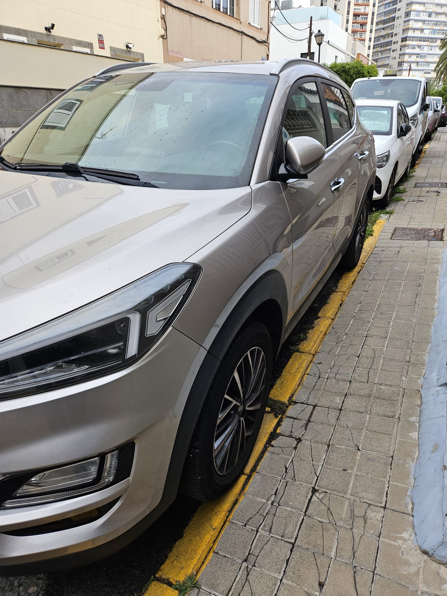 SUV plateada estacionada en la acera, con detalles visibles de la carrocería y llantas. Al fondo, se aprecian otros vehículos y edificios en un entorno urbano.