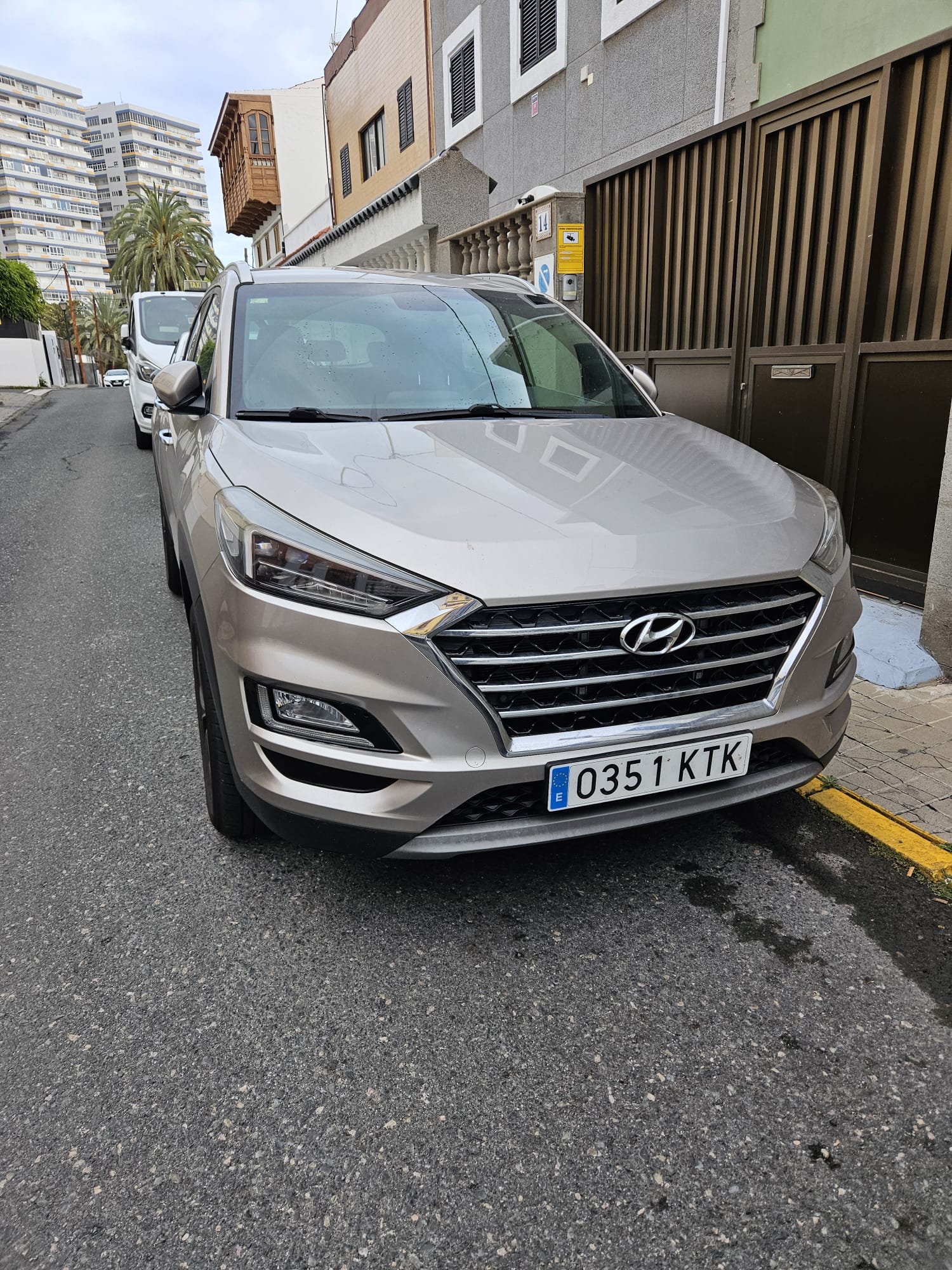 SUV Hyundai estacionada en una calle urbana, con edificios residenciales y palmeras al fondo. La imagen muestra el diseño frontal del vehículo, incluyendo su parrilla distintiva y matrícula.