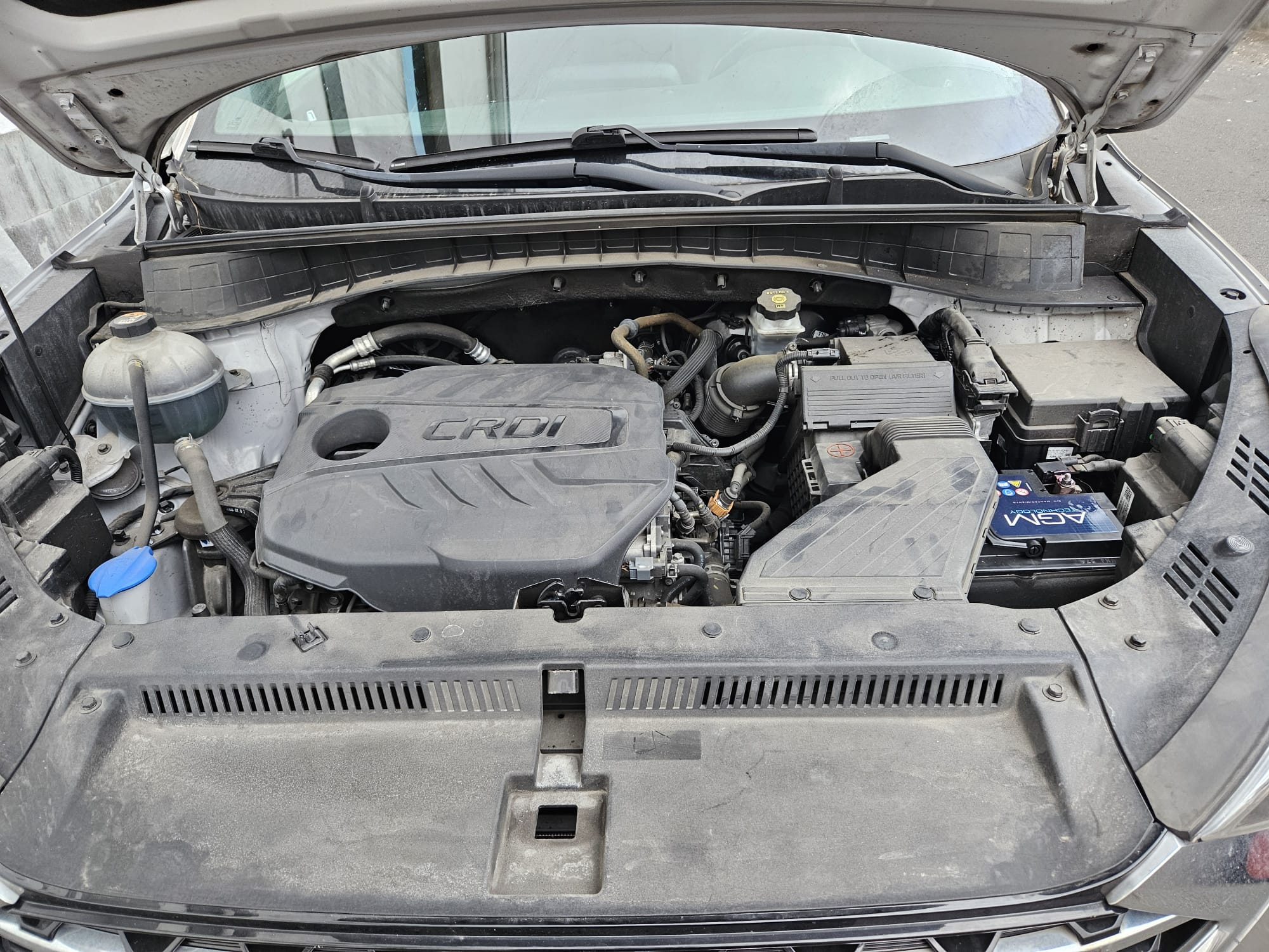 Imagen del motor de un vehículo con el capó abierto, mostrando el sistema CRDi, componentes eléctricos y un acumulador, todo con una capa de polvo visible.