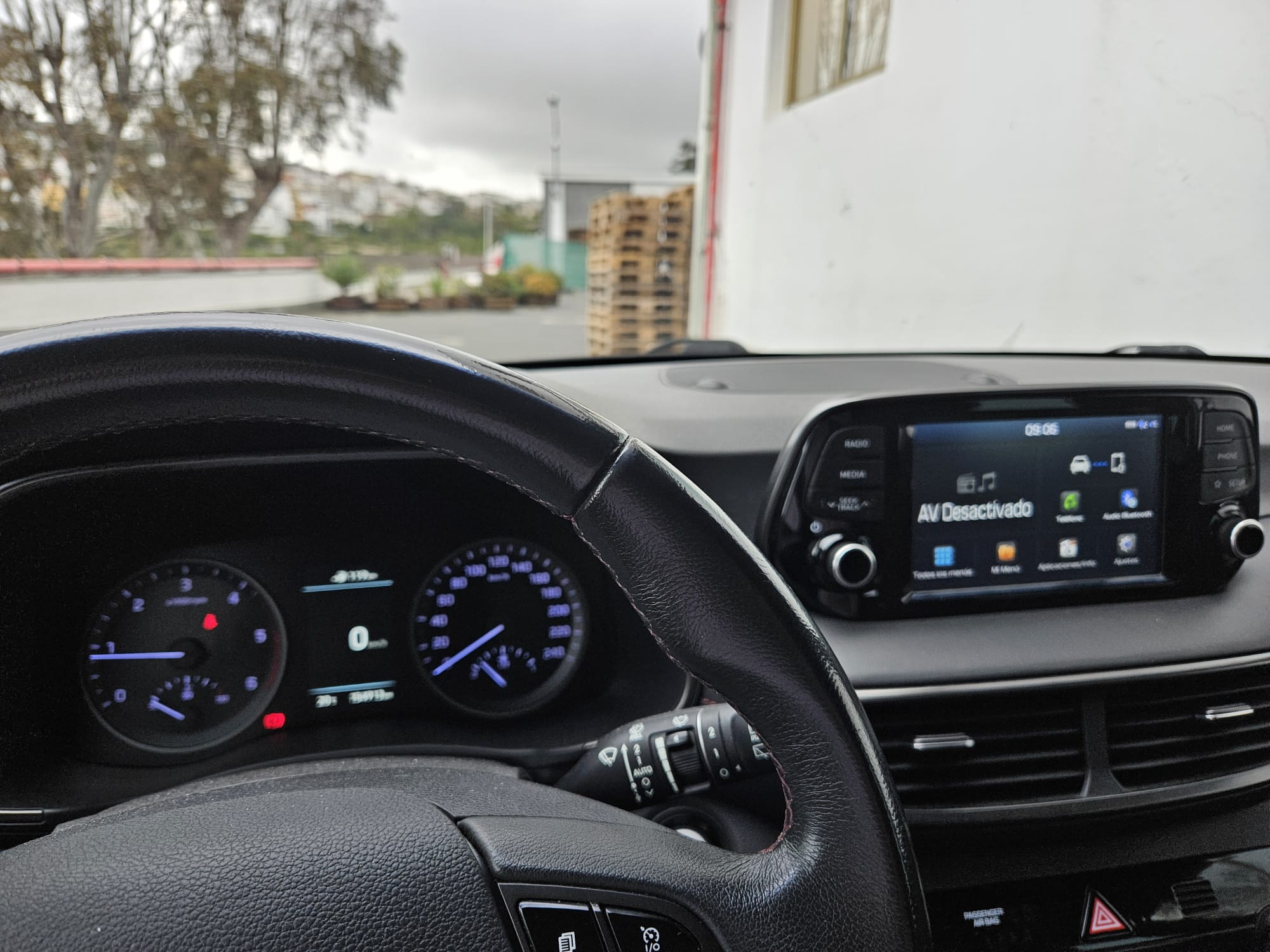 Interior de un coche mostrando el volante y el tablero de instrumentos, con el sistema de infoentretenimiento encendido. Se pueden ver indicadores de velocidad y combustible, así como un mensaje que indica "AV Desactivado". En el fondo, se observa un paisaje urbano y palets de madera.