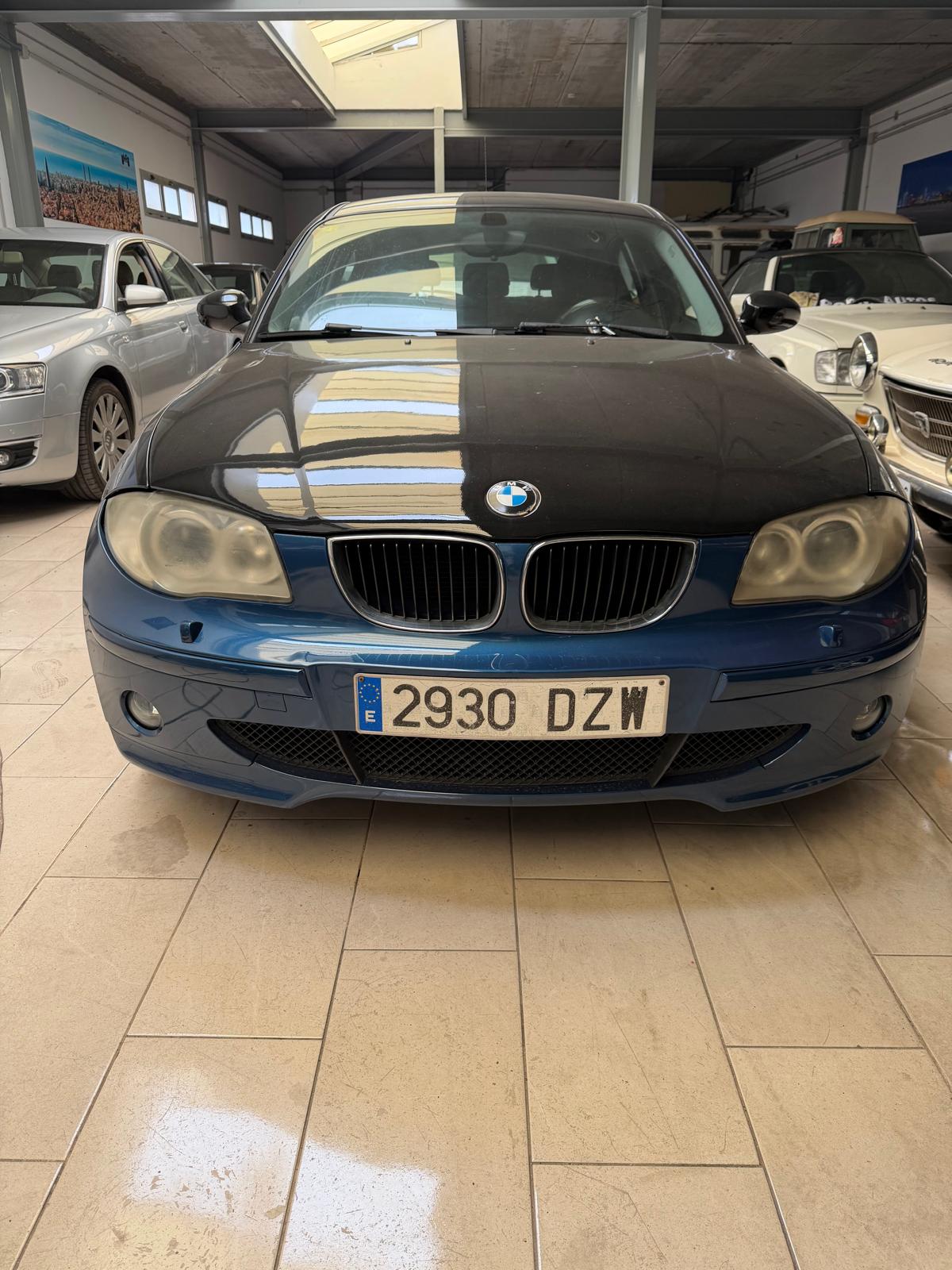 BMW serie 1 estacionado en un garaje, con carrocería negra y azul, y matrícula 2930 DZW. Al fondo, se pueden ver otros vehículos y un ambiente luminoso.