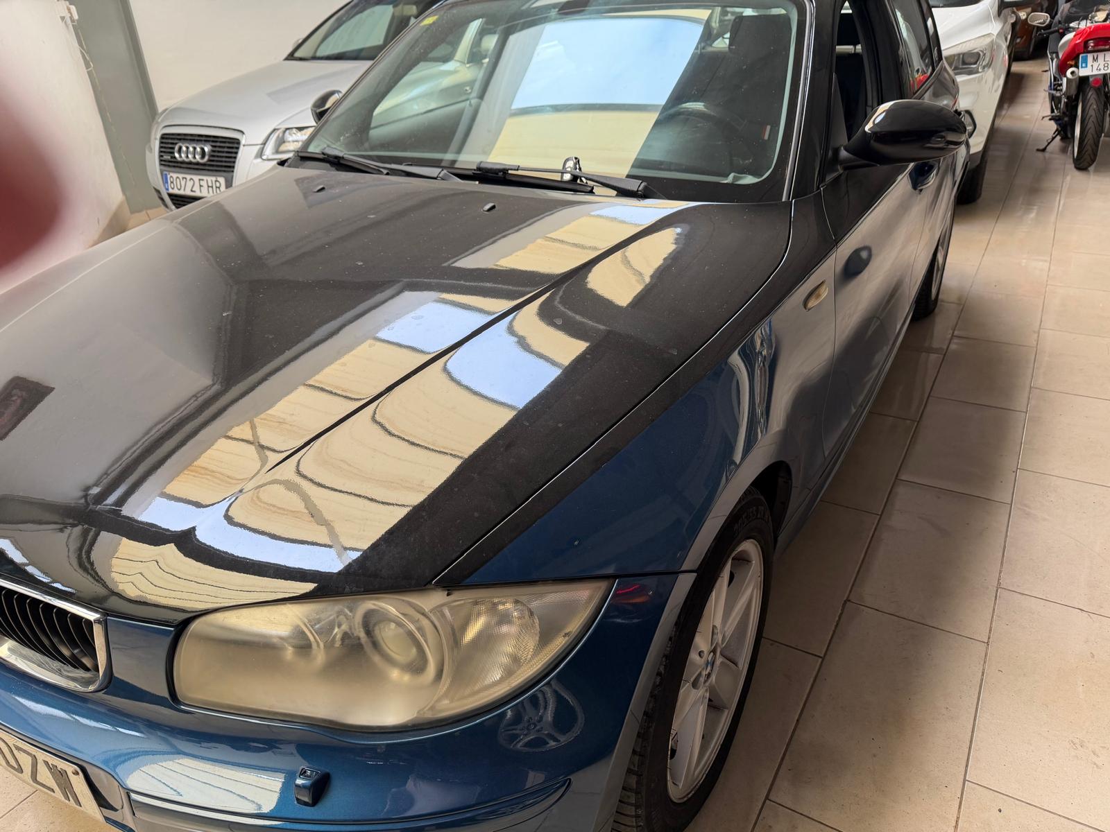 Automóvil BMW de color azul con capó negro, estacionado en un garaje. Se observa una superficie polvorienta y reflejos de luz en el capó. Otros vehículos están aparcados en el fondo.
