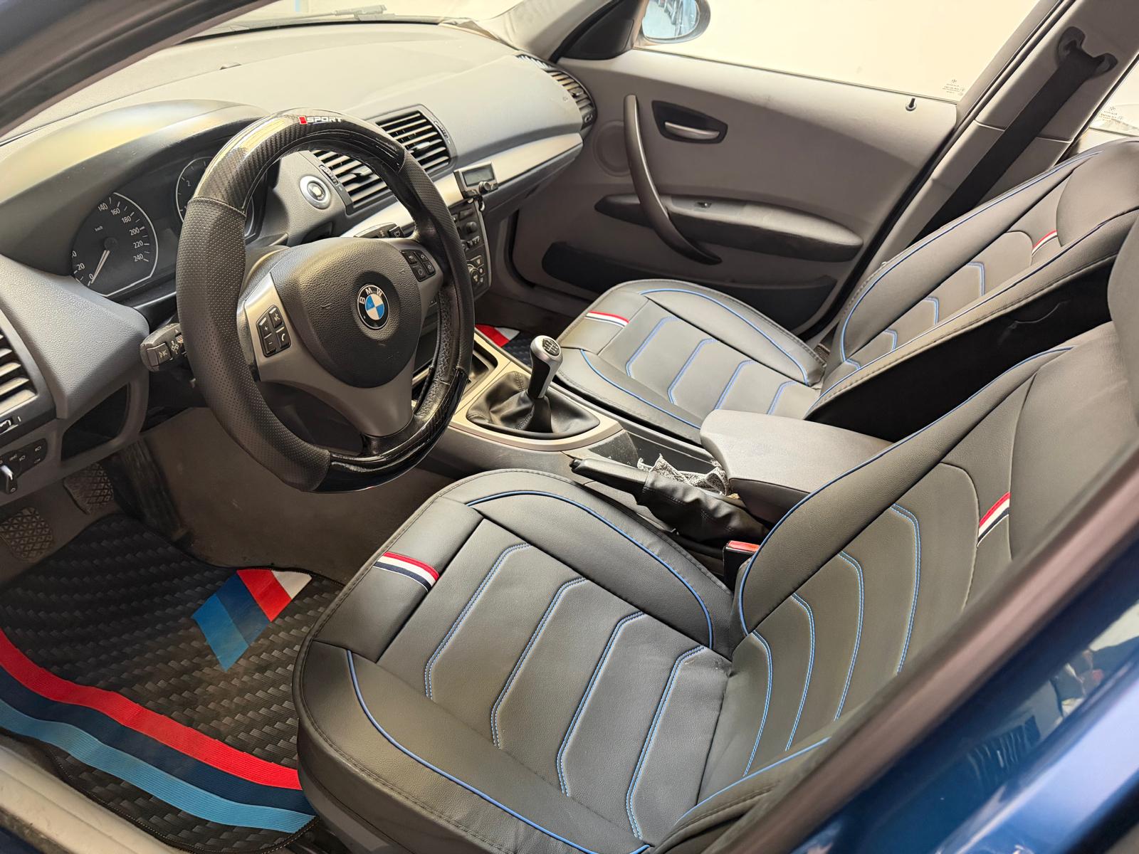 Interior de un automóvil BMW, mostrando un volante deportivo, asientos de cuero negro con detalles en azul y rojo, y un tapete con el logo M. La cabina destaca por su diseño moderno y funcional.