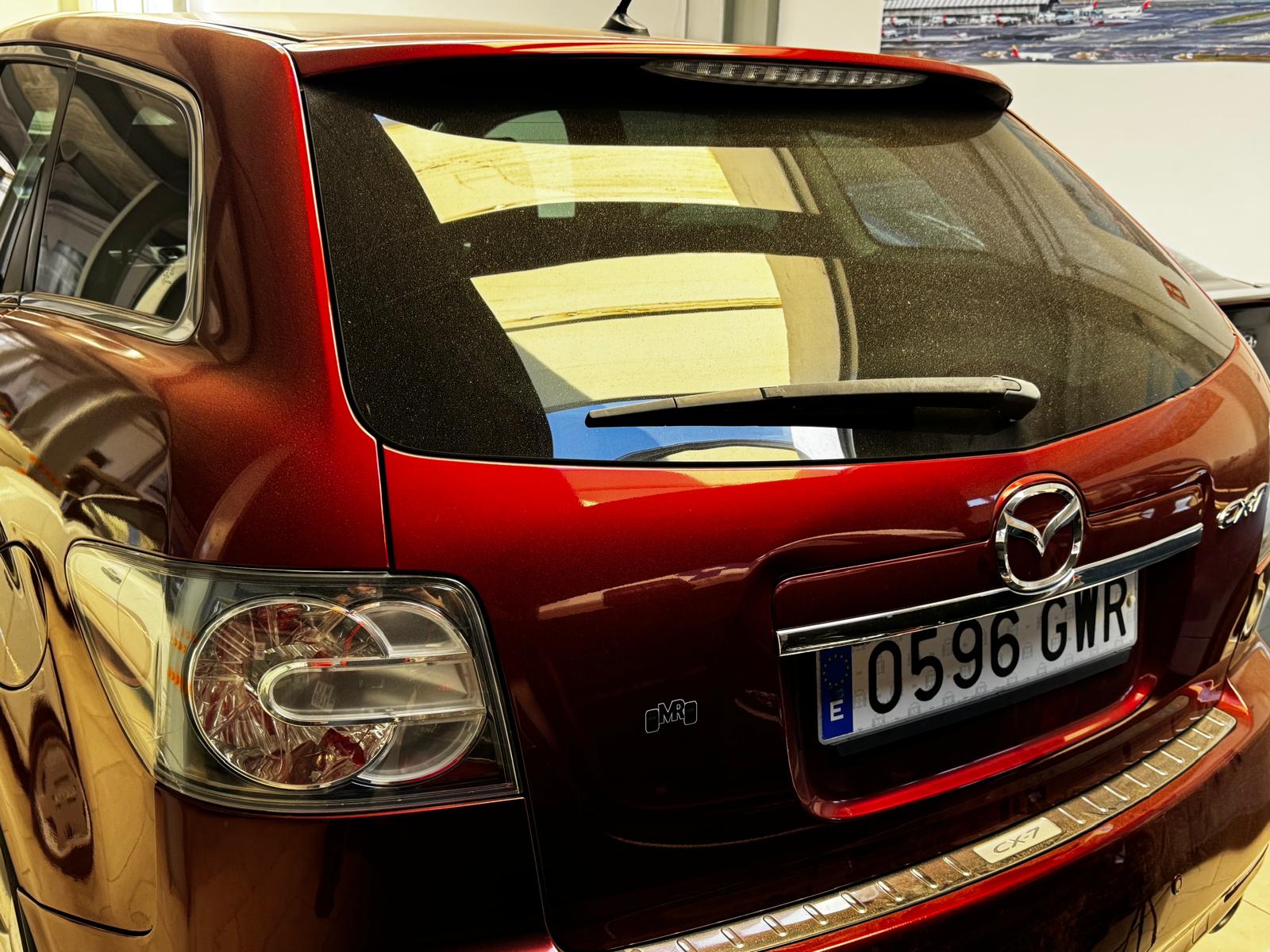 Vista trasera de un automóvil Mazda CX-7 de color rojo, mostrando detalles del diseño, luces traseras y matrícula con el número 0596 GWR. El vehículo se encuentra en un entorno cerrado, evidenciando un acabado brillante y limpio.
