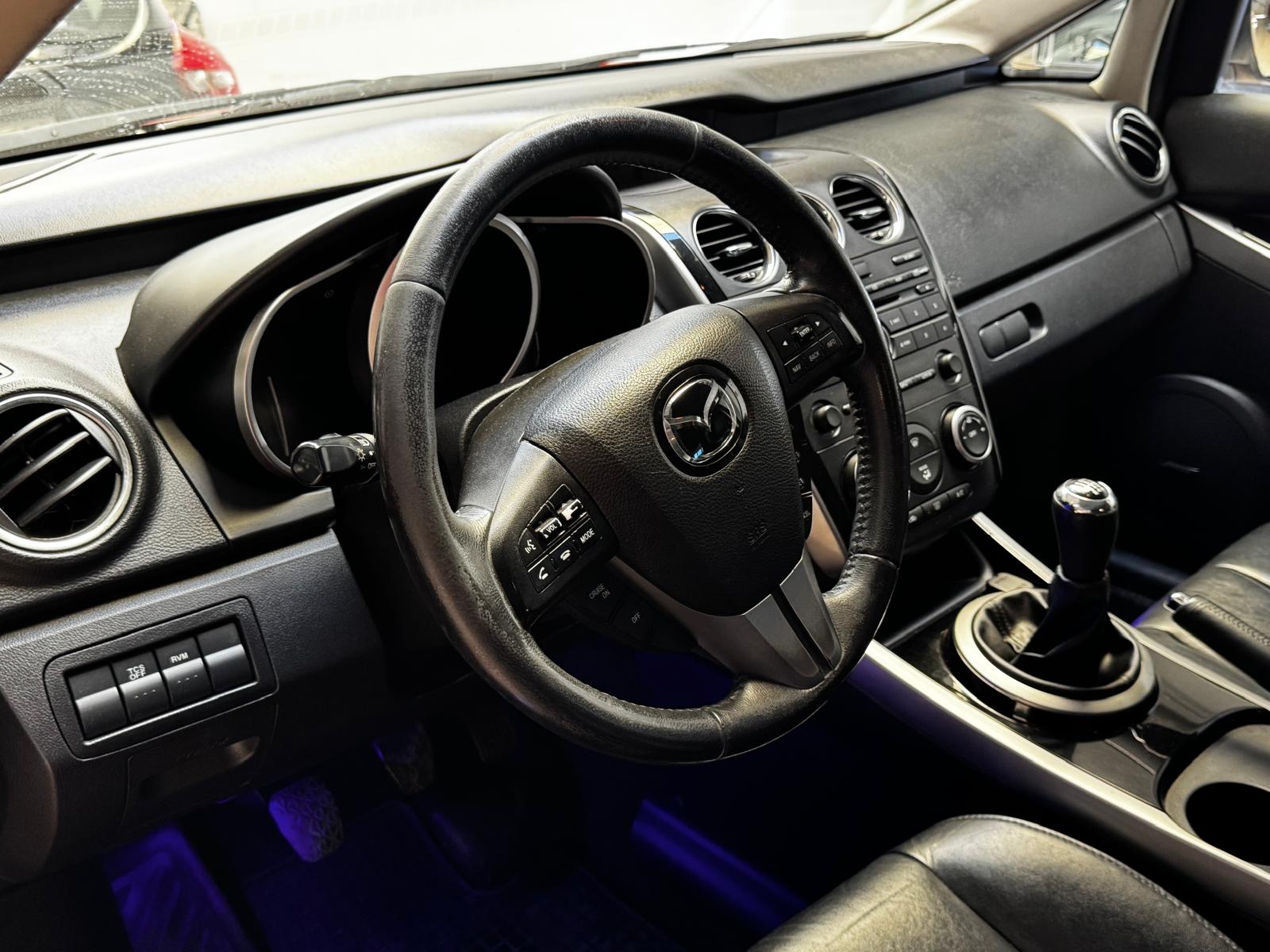 Interior de un automóvil Mazda con volante de cuero, tablero moderno, controles de audio y aire acondicionado. Se observa un cambio de marchas manual y asientos de cuero. Iluminación ambiental azul en el suelo.