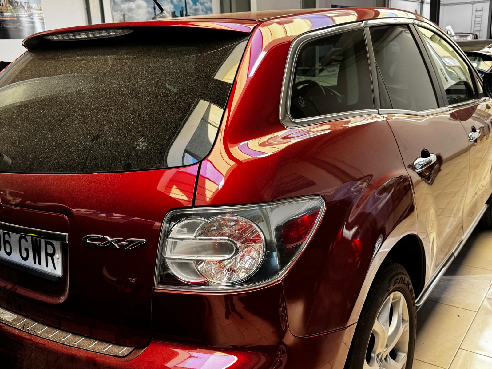 SUV Mazda CX-7 roja estacionada en un concesionario, mostrando su diseño moderno y detalles en la carrocería. La parte trasera presenta un vidrio ligeramente sucio y luces traseras bien definidas.