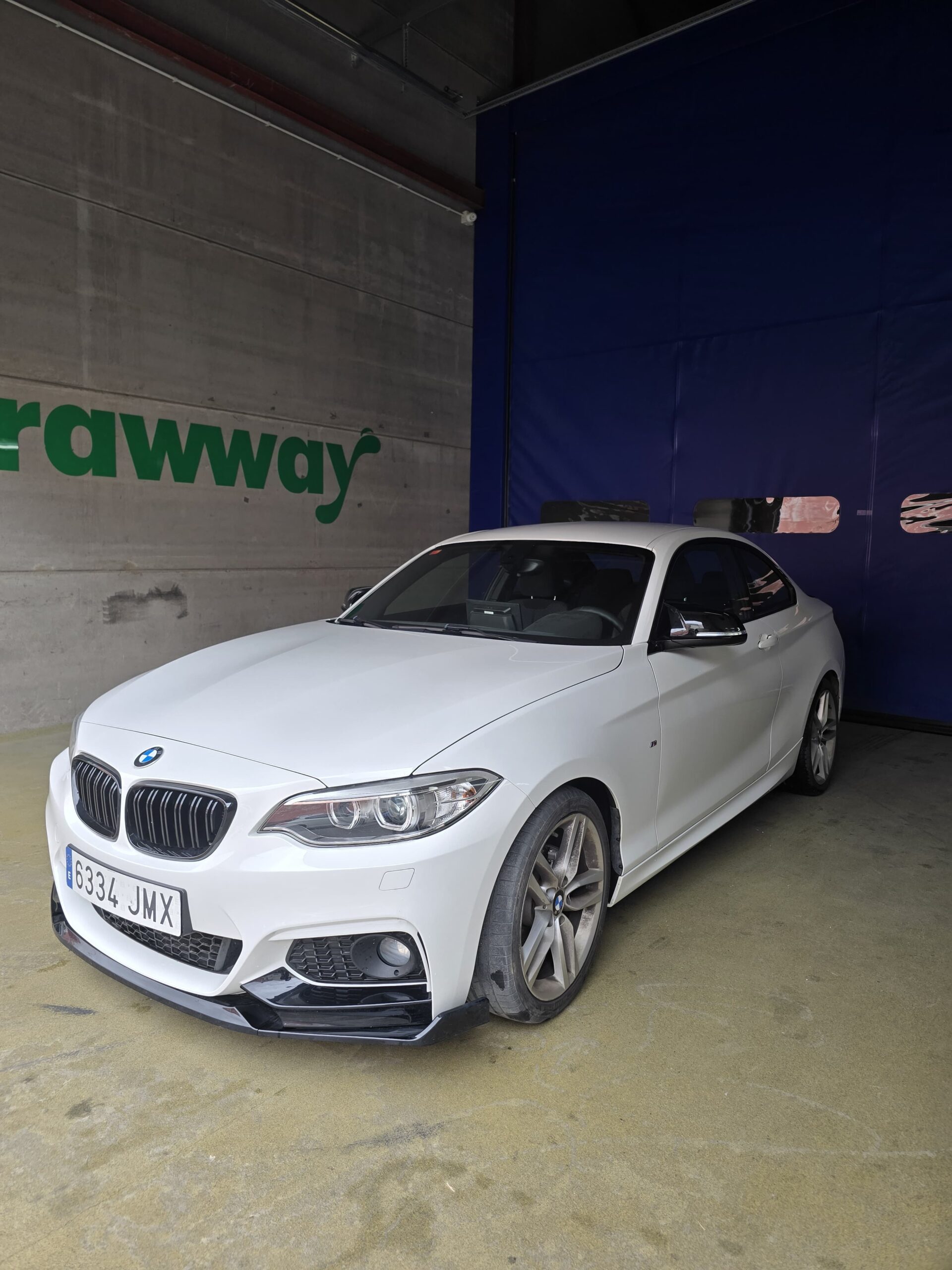 Automóvil BMW blanco estacionado en un garaje, con un diseño deportivo y llantas de aleación. El fondo muestra una pared con el logo "rawway".