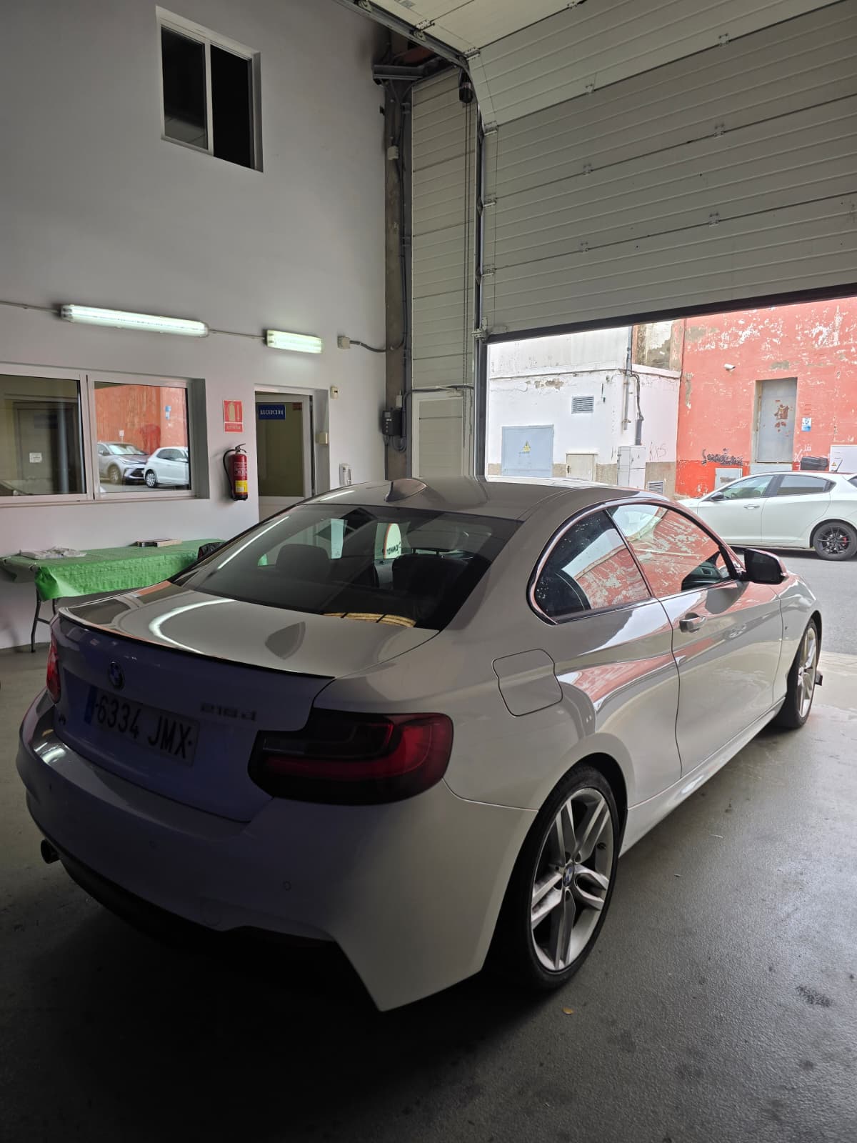 Coupé BMW blanco estacionado en un taller, con luz natural entrando por la puerta abierta, mostrando el interior y la pared de fondo.