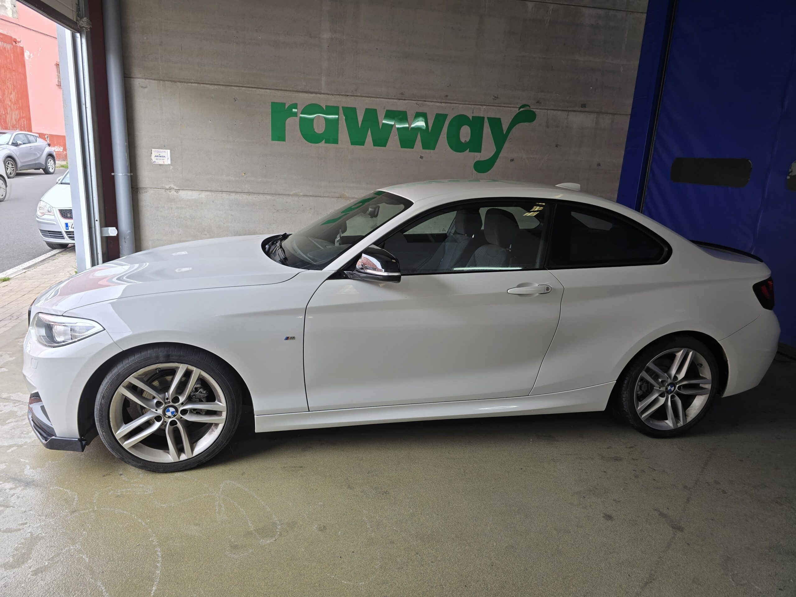 Coche deportivo blanco BMW serie 2 estacionado en un taller, con el logo visible y el letrero "rawway" en la pared.