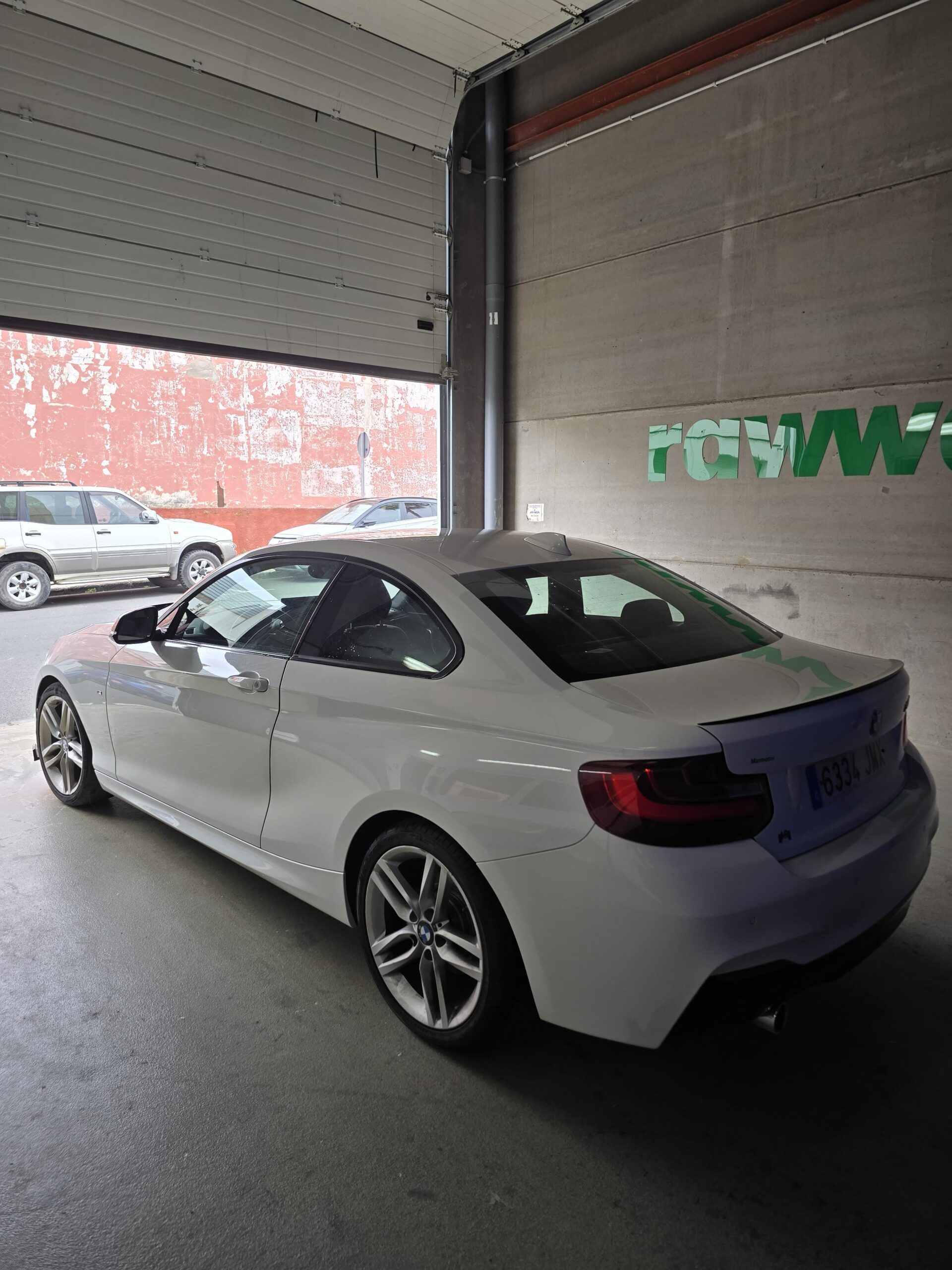 Coche deportivo blanco BMW estacionado en un garaje con pared de ladrillo rojo al fondo. Se pueden ver las llantas de aleación y el diseño aerodinámico del vehículo.